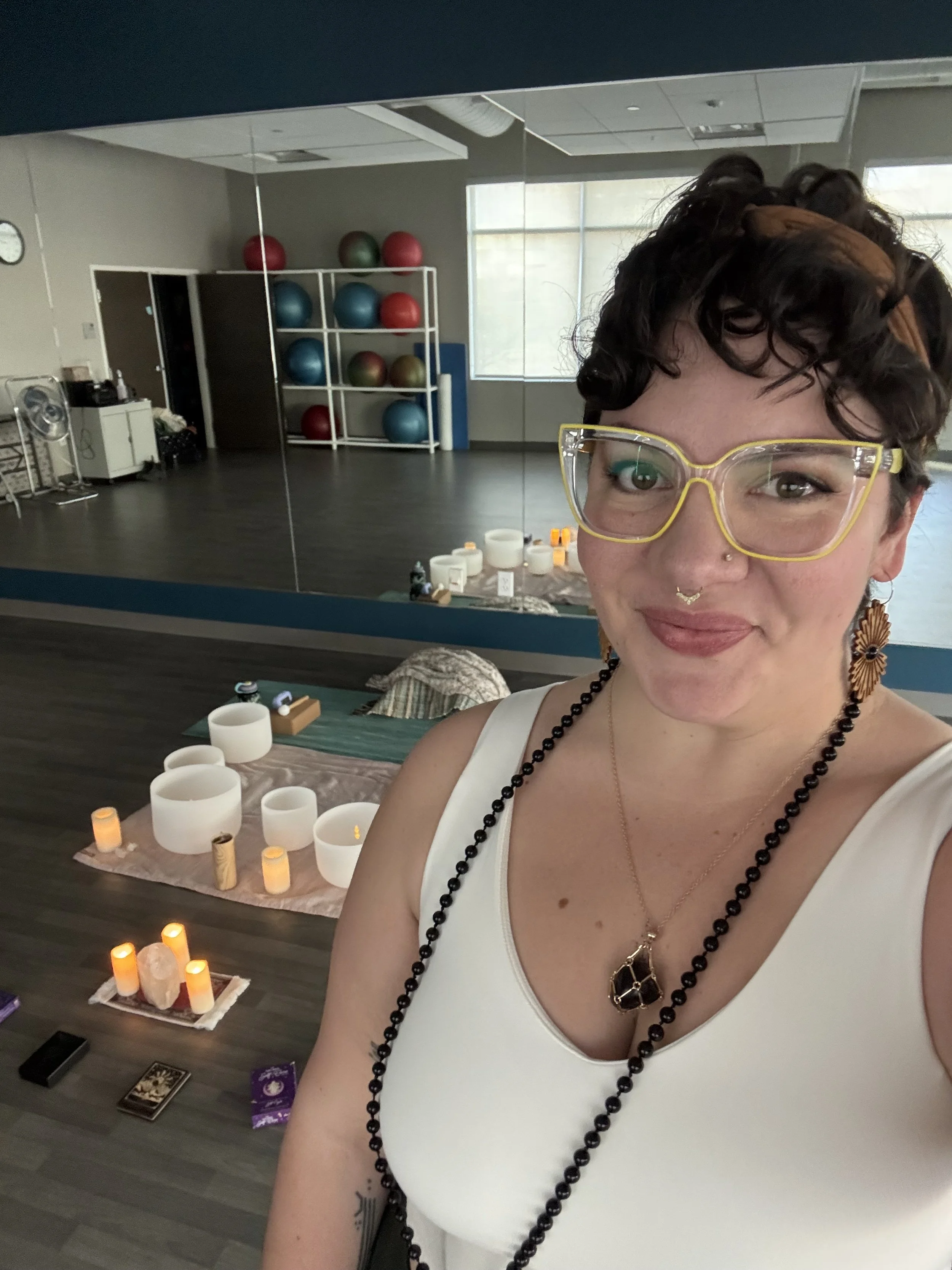 A woman with curly dark hair, yellow glasses, and hoop earrings takes a selfie in a yoga or meditation studio. The studio has a mirror, shelves with exercise balls, and a table with candles, salt lamps, and crystals.