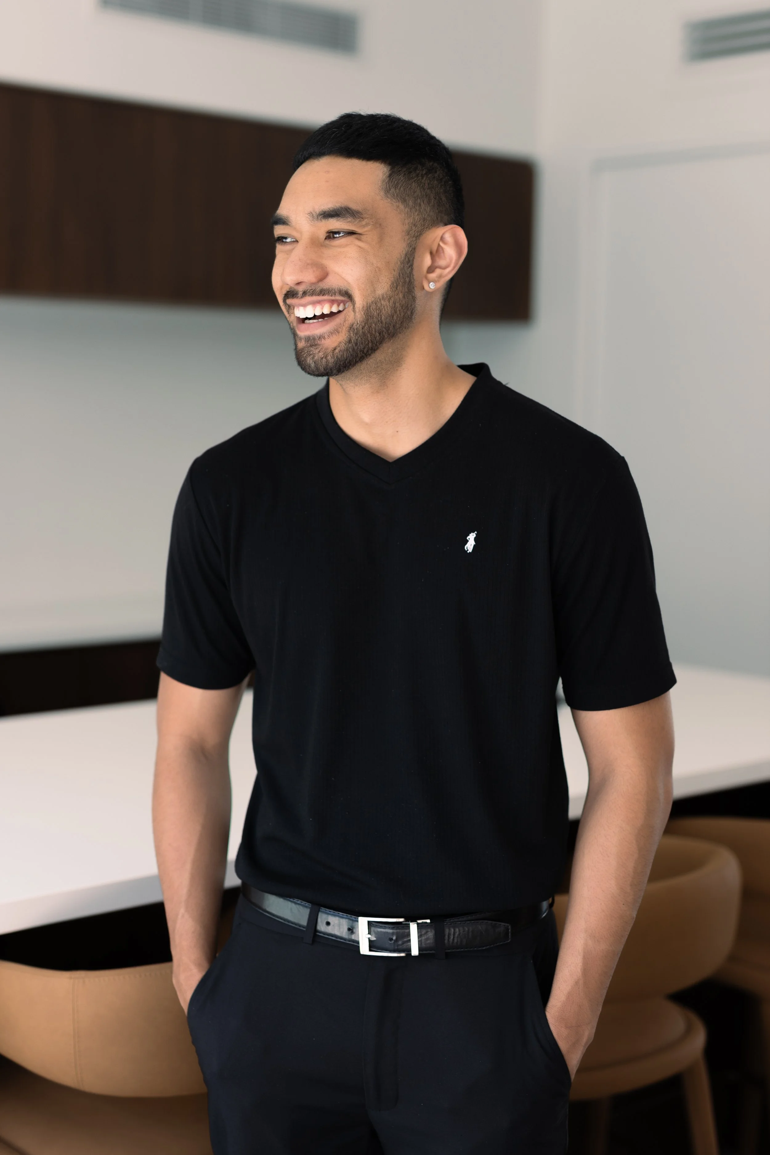 A young man with short dark hair, a beard, and earrings, smiling and wearing a black t-shirt and black pants, standing in a modern indoor setting.