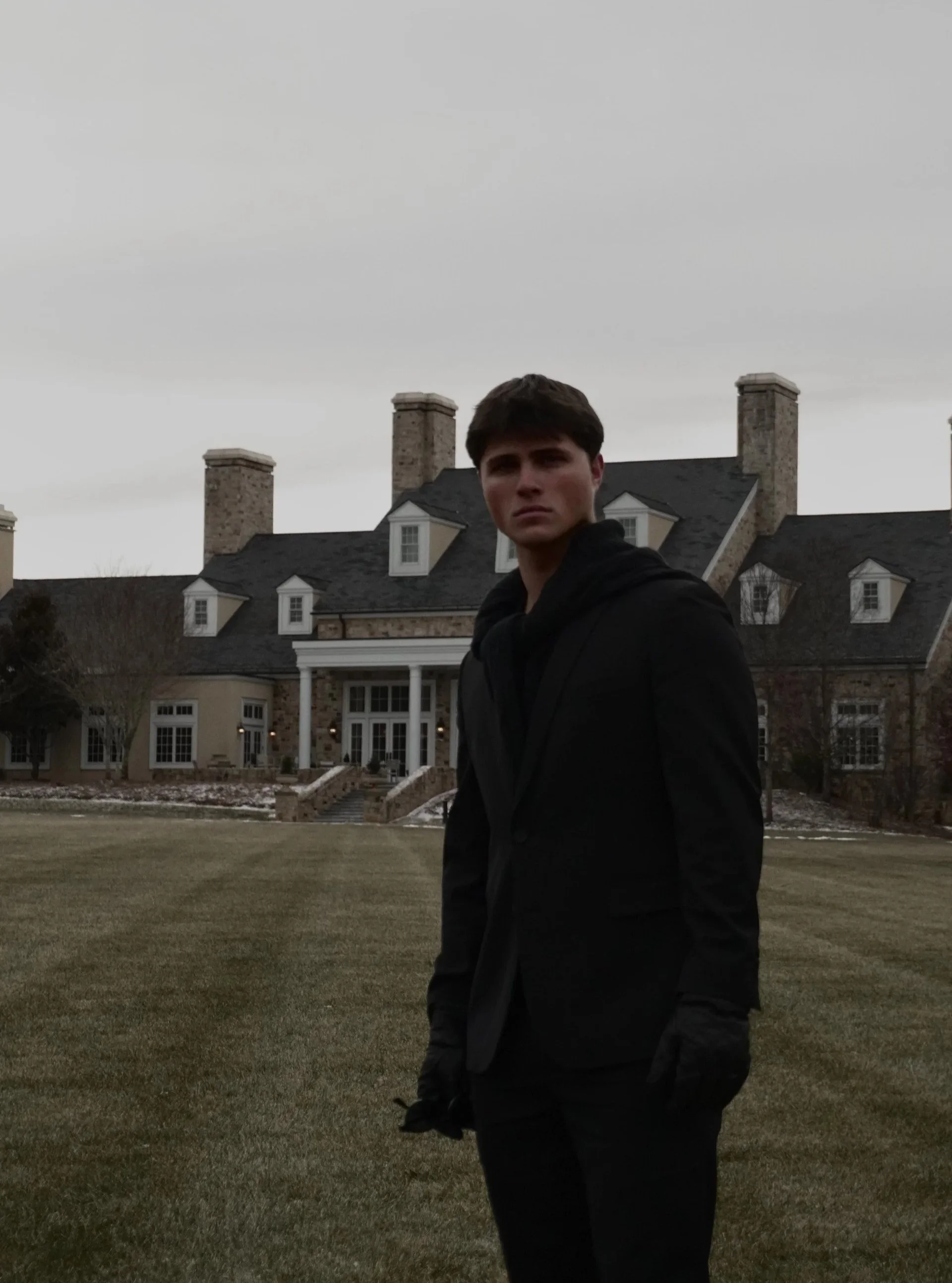 A young man in a black suit and gloves stands on a lawn in front of a large mansion on a cloudy day.