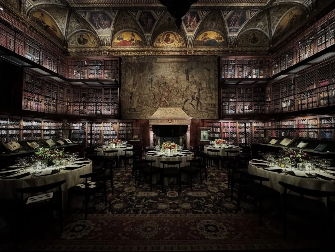 Elegant library room set up for a formal event, with round tables covered in white tablecloths, floral centerpieces, chairs, and surrounded by floor-to-ceiling bookshelves behind decorative ironwork. The room features ornate ceiling murals, a large tapestry on the wall, and a fireplace.