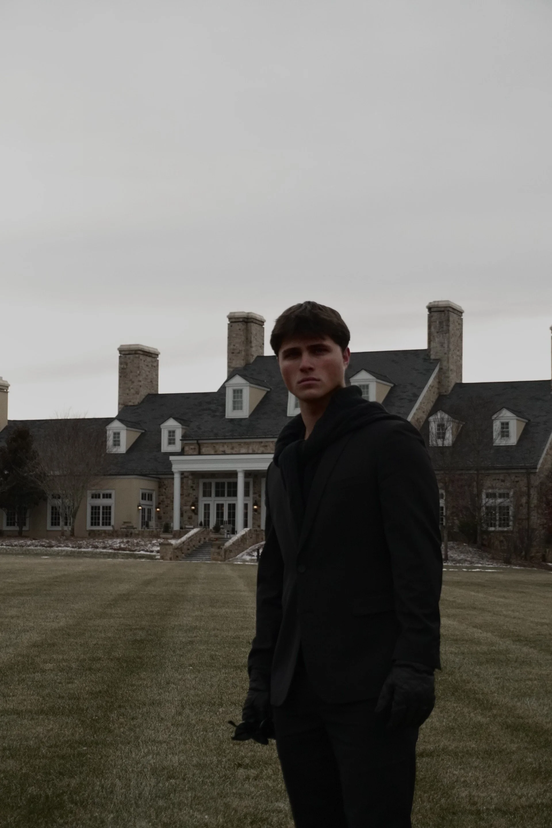 A young man in black clothing standing on a lawn in front of a large, historic-looking house under an overcast sky. This man is the Founder of Aston Thomas, Evan Carmello
