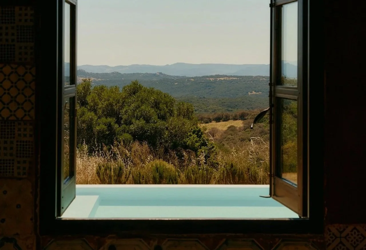 Open window showing a hillside landscape with trees, fields, and distant mountains under a clear sky.