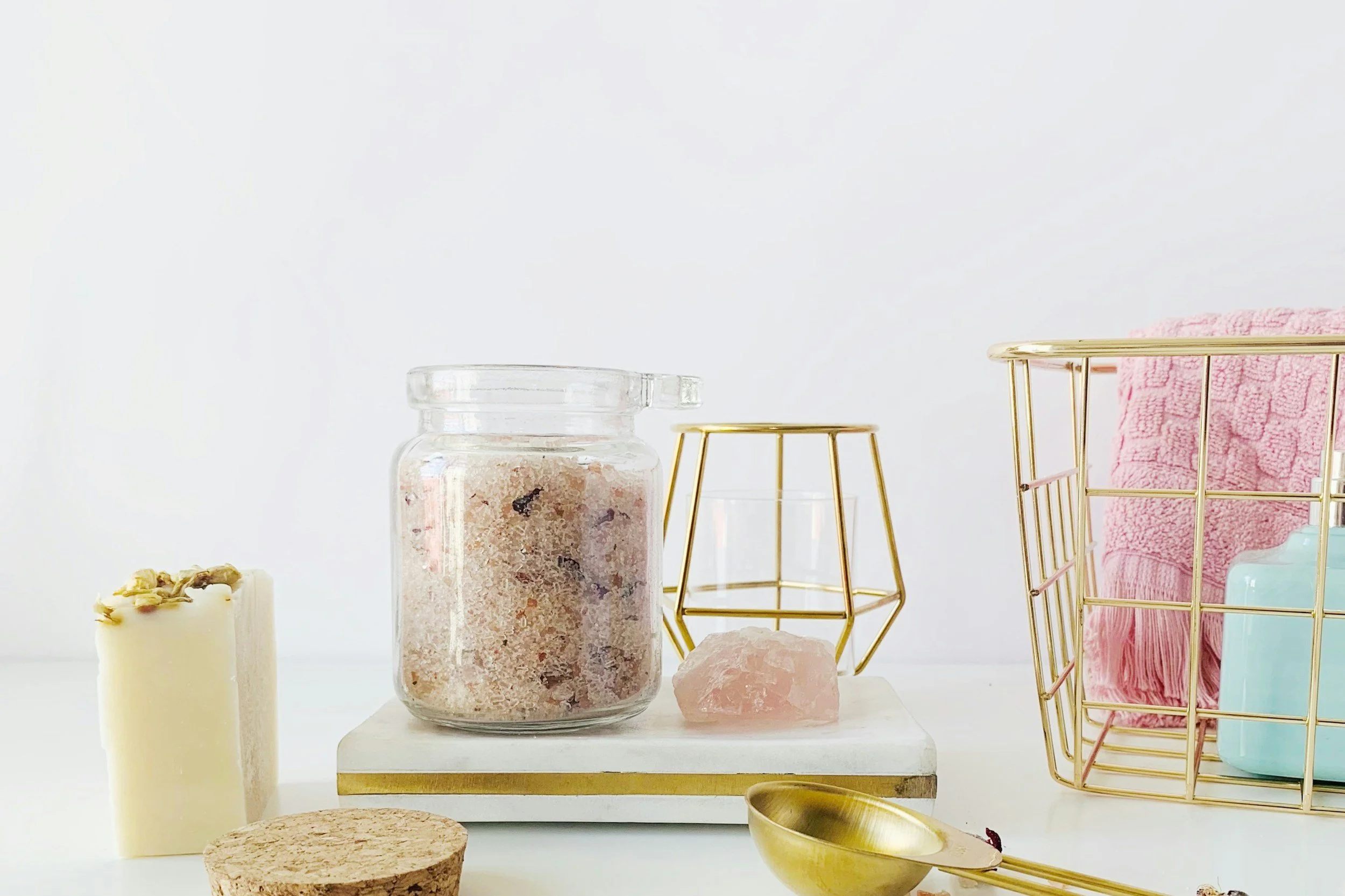 Bathroom or wellness decor with candles, a pink Himalayan salt crystal, a gold wire basket, and a pink towel on white surface at The one beauty bar in San Antonio