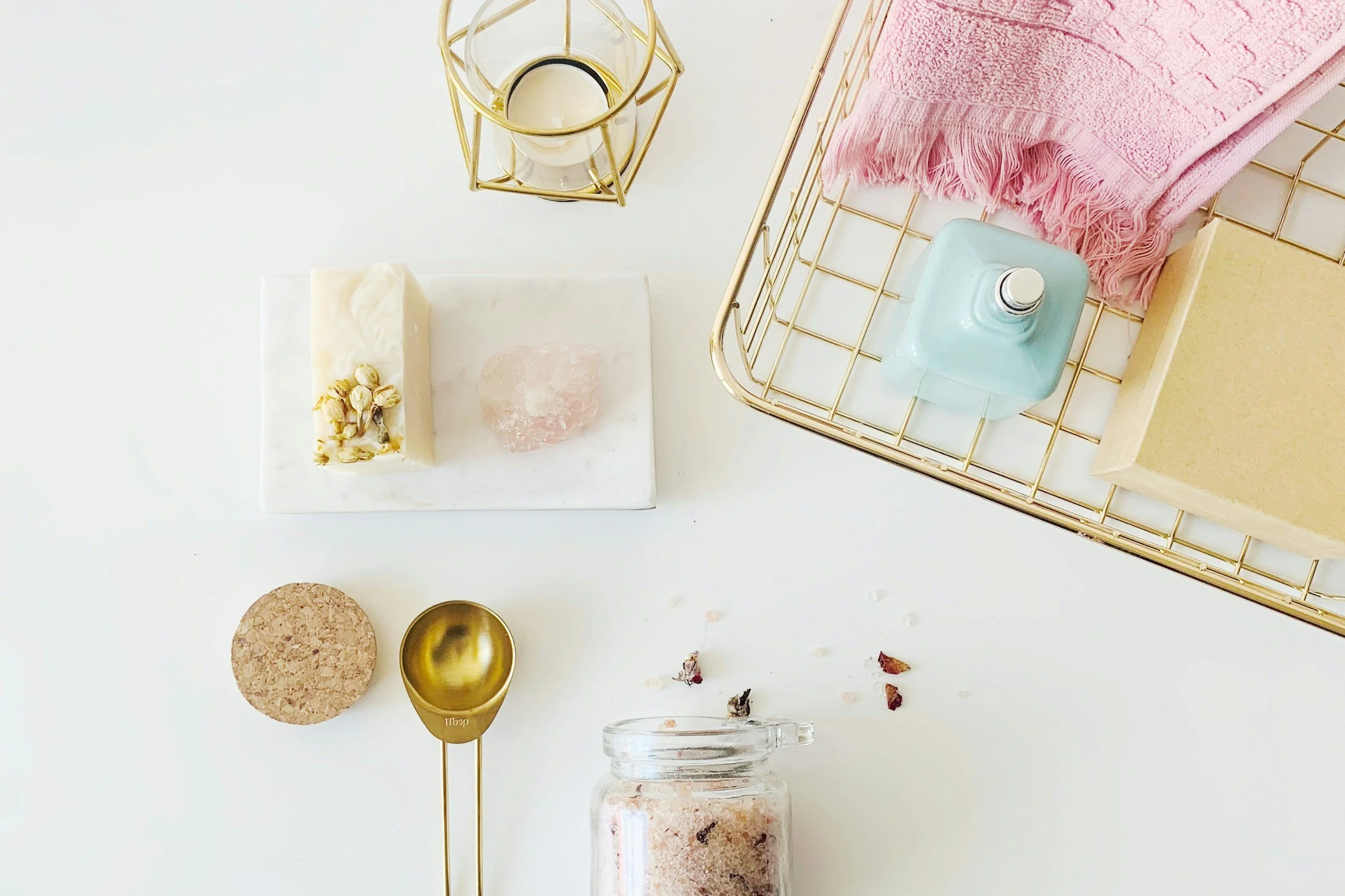 Flat lay of bathroom accessories on a white surface, including a soap bar with dried flowers, a pink salt rock, a round cork coaster, a gold spoon, a glass jar of bath salts, a gold wire basket with a pink towel and a light blue bottle, and a gold candle holder with a white candle.