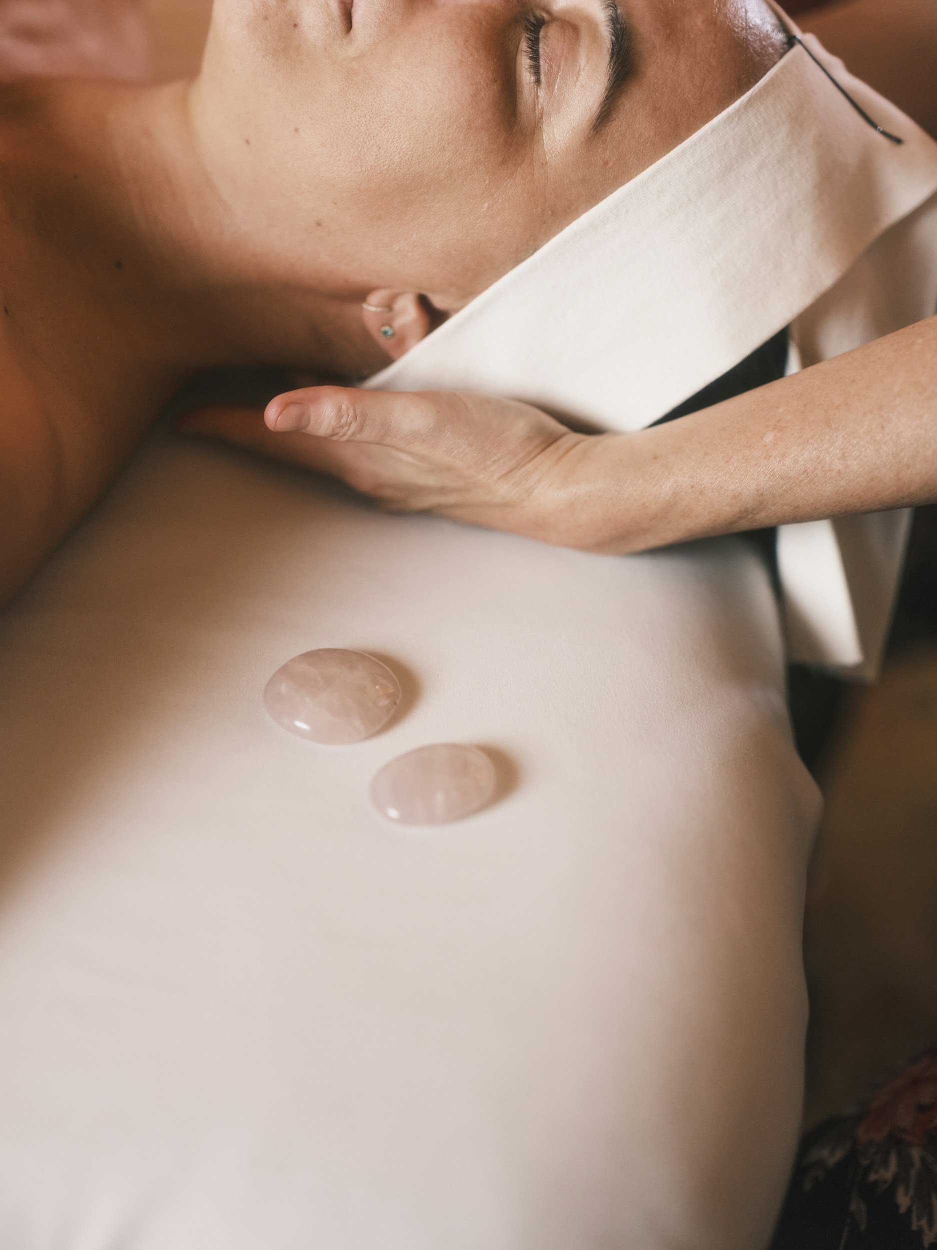 A woman receives a facial massage during a spa treatment, with two seashell-shaped stones resting on her shoulder.