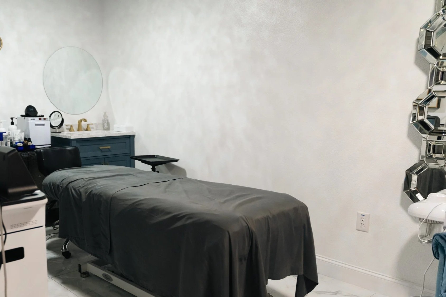 A medical examination room with a massage table covered in black sheets, a blue cabinet with a sink and mirror, various medical supplies, a black chair, and chrome hexagon-shaped wall mirrors.