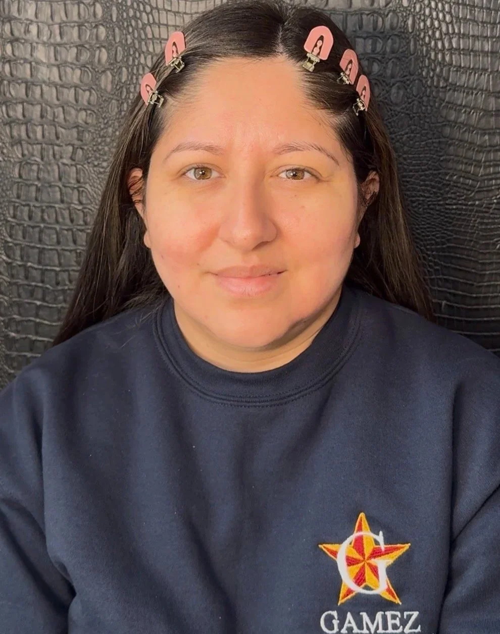 A woman with long dark hair clipped with pink hair clips, sitting against a textured black background, wearing a navy sweatshirt with a logo and the word "GAMEZ" on it.