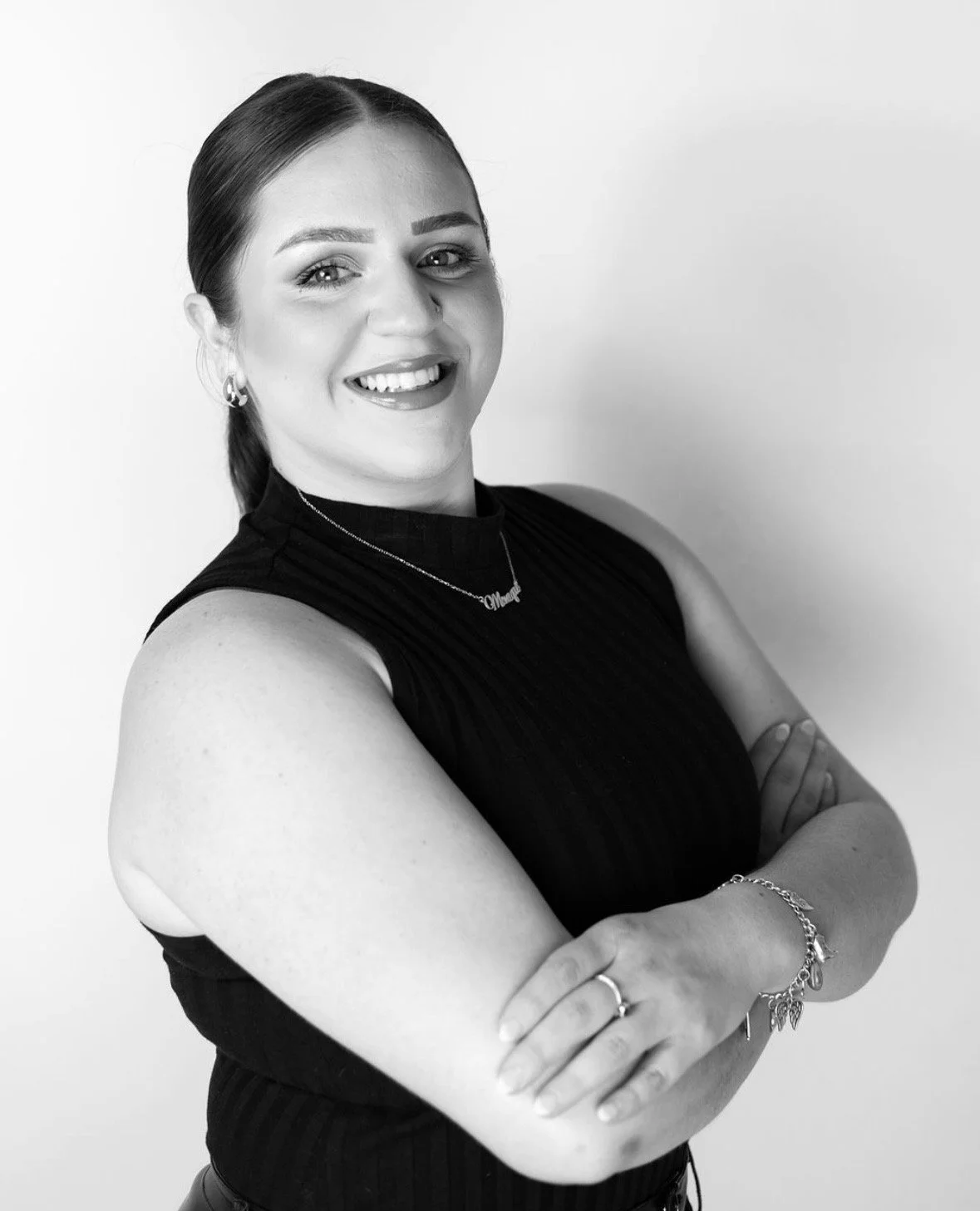 A woman smiling with arms crossed, wearing a sleeveless black top and jewelry, standing against a plain background.