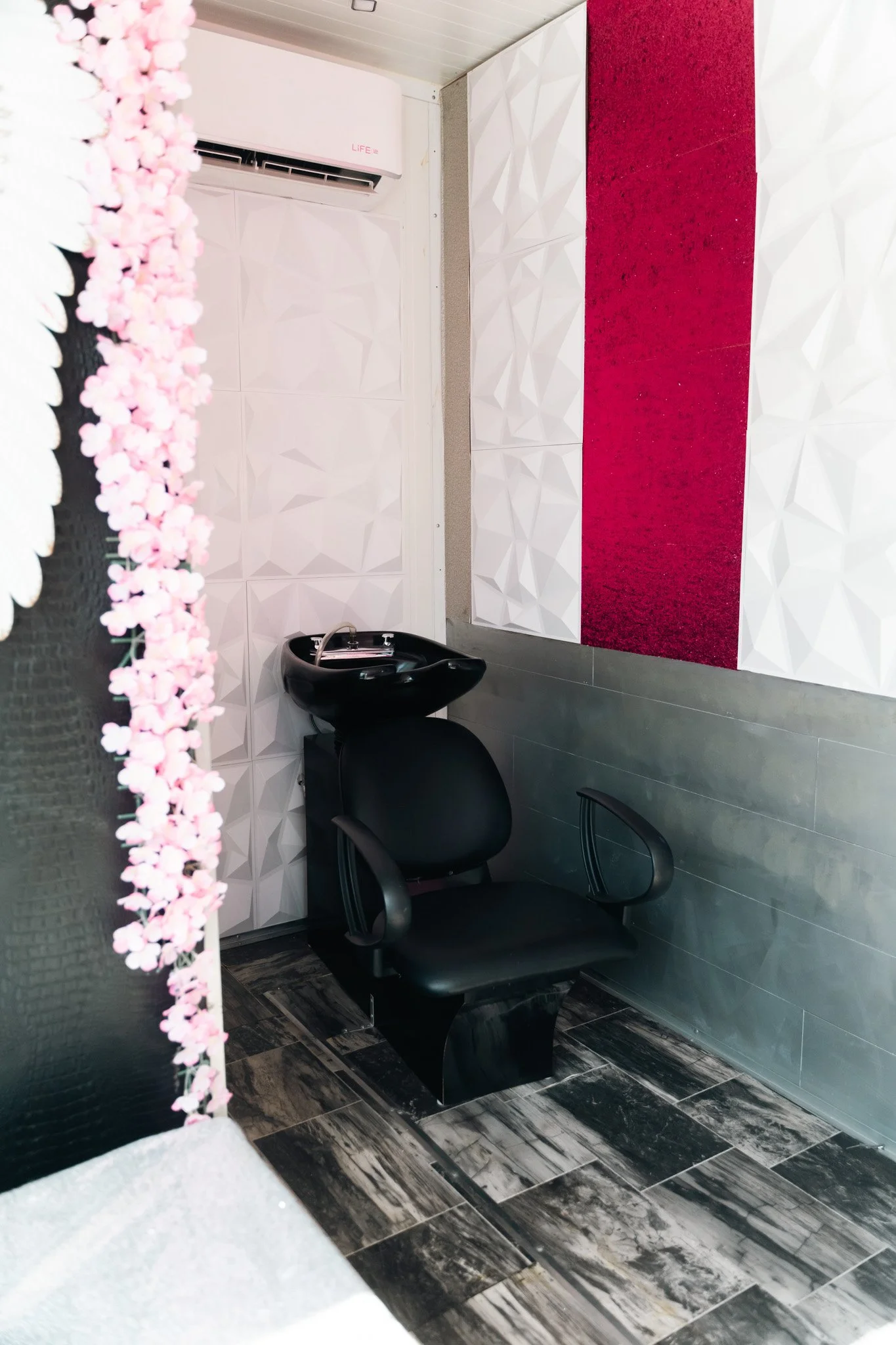 A small salon or barber shop wash area with a black shampoo sink, a black chair, decorative textured white and red walls, black and gray tiled floor, and an air conditioning unit.