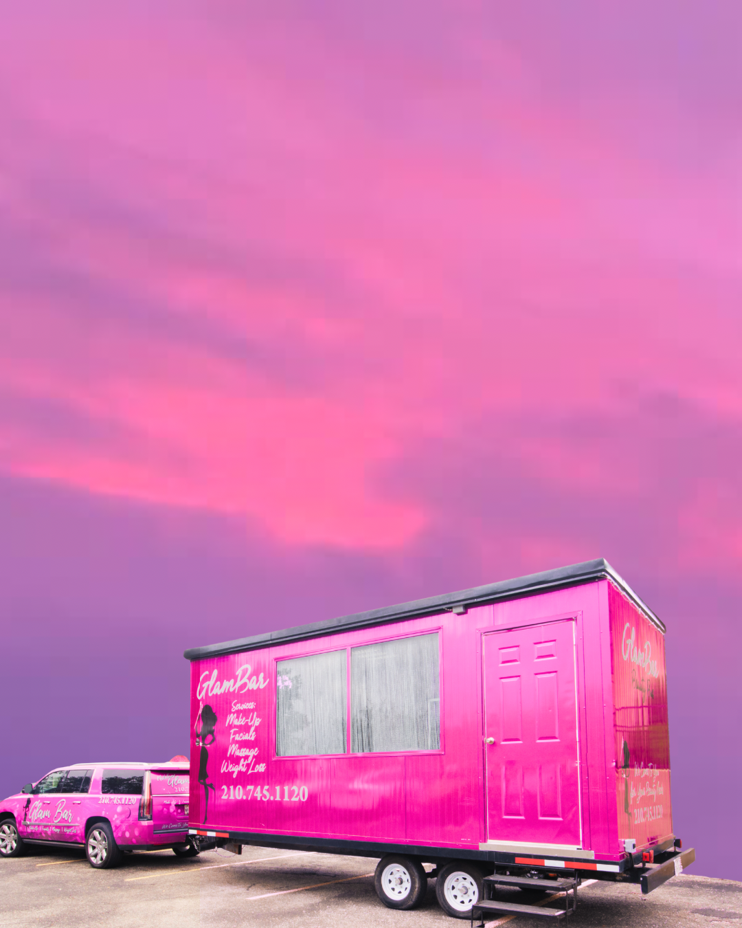Pink mobile beauty salon trailer and car parked on an empty lot against a pink and purple sunset sky.
