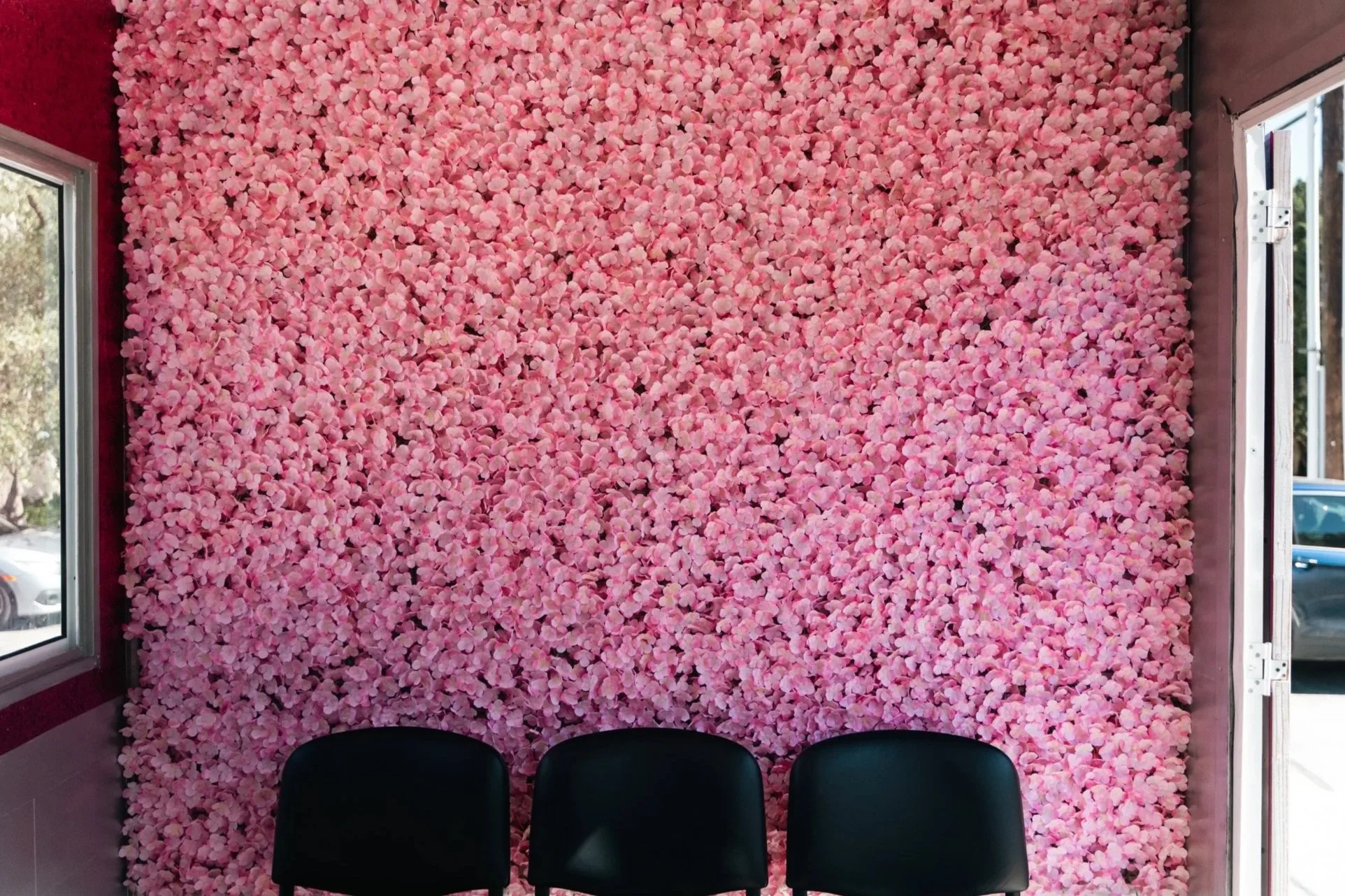 Pink flower wall with three black chairs in front of it, inside a room with a window on the left and open door on the right.