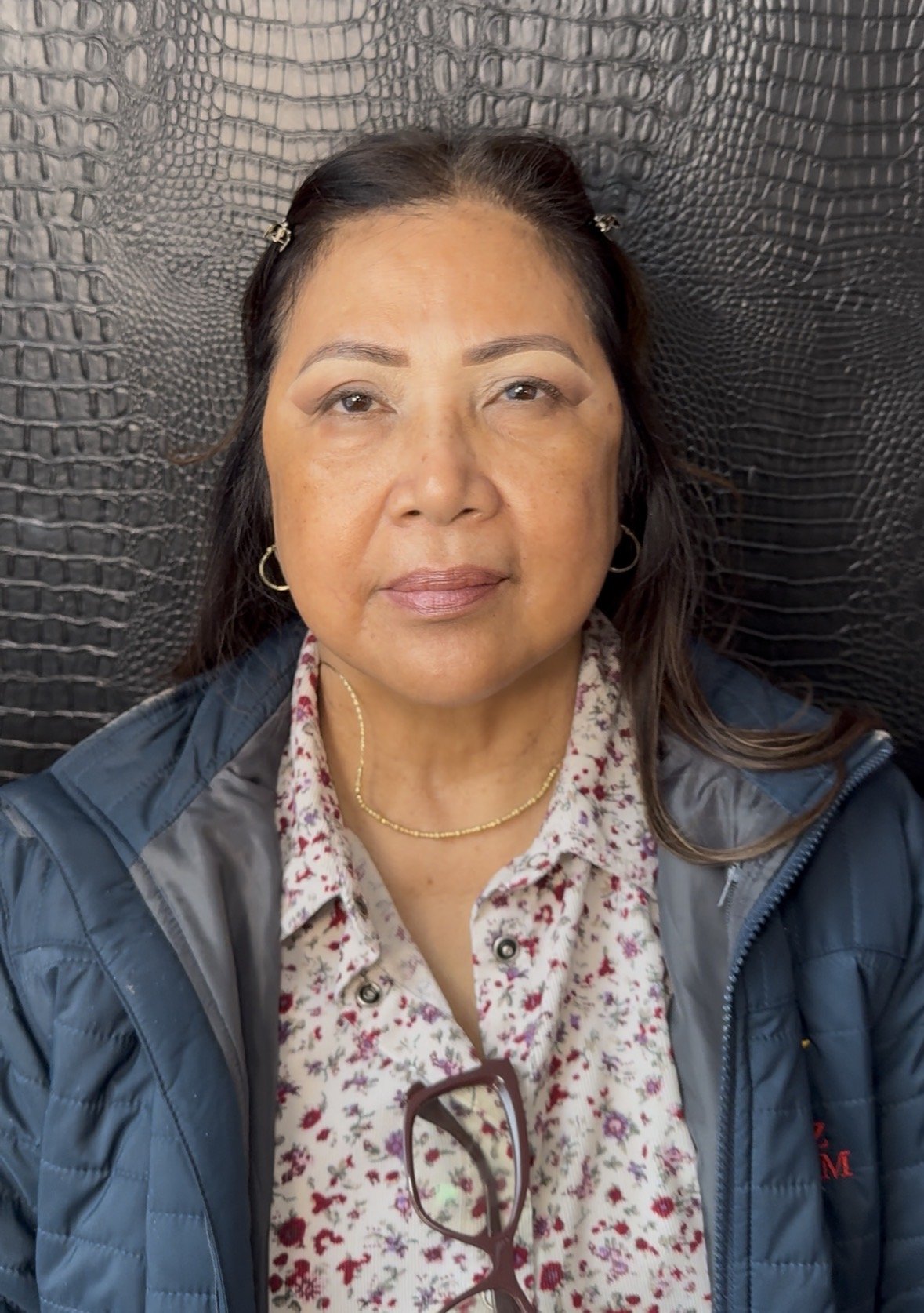 A middle-aged woman with dark brown hair and light makeup, wearing a floral blouse, gold jewelry, a blue jacket, and glasses hanging from her collar, sitting in front of a black textured background.