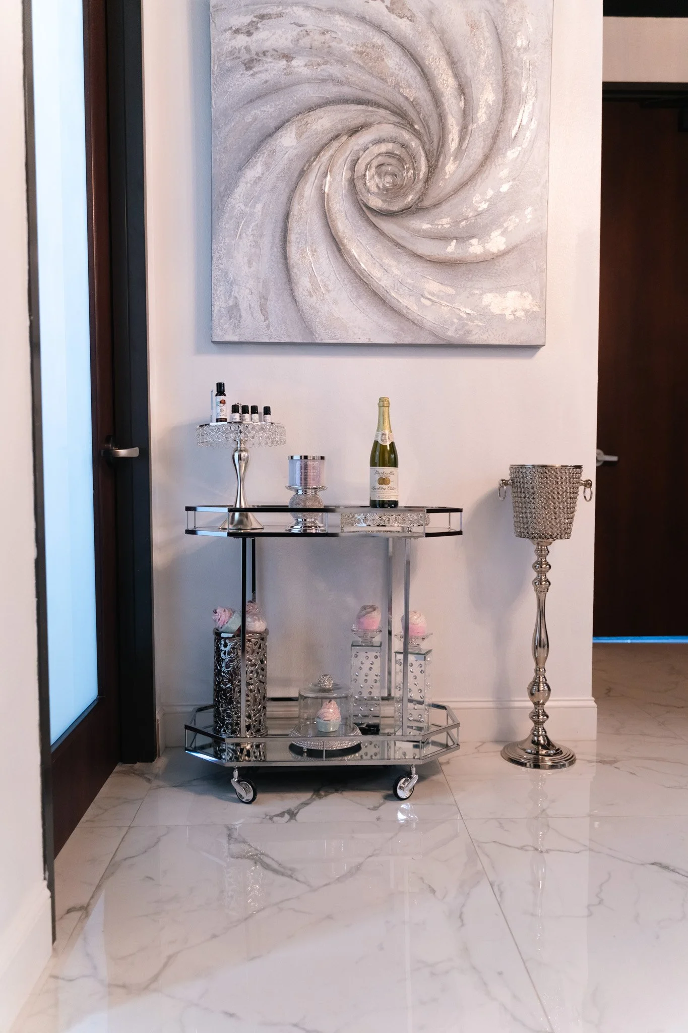 A decorative cart with pink cupcakes, a champagne bottle, and makeup items, positioned in front of a white wall with a large abstract swirl painting, standing beside a silver pedestal with a metallic lamp on it.