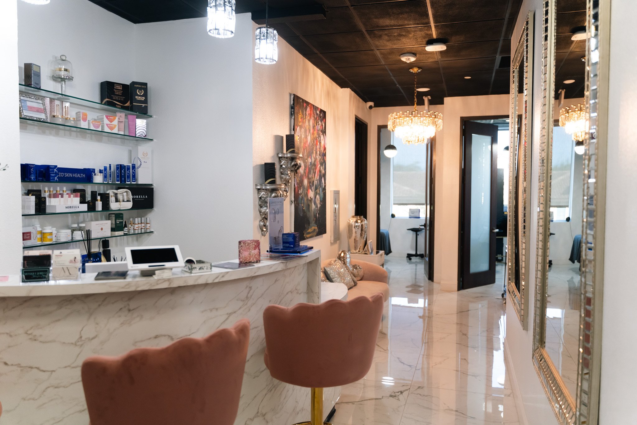 Interior of a modern spa or beauty clinic with a marble reception desk, pink velvet chairs, decorative mirrors, and a hallway with chandeliers.