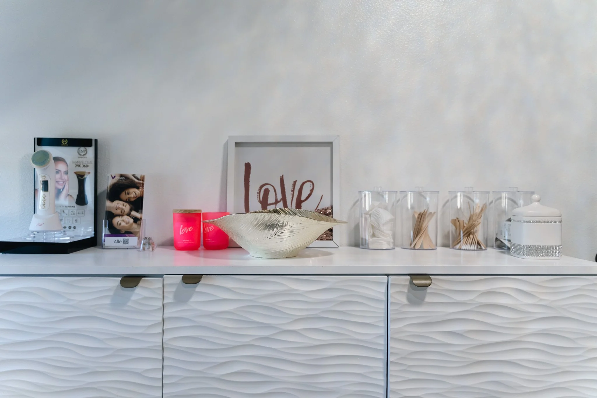 White sideboard with textured doors decorated with a framed love quote, a ceramic bowl, pink candles, a hand-held beauty device, toothpicks, wooden utensils, and a jewelry box, with a wall-mounted mirror.