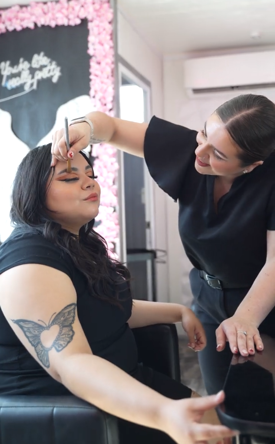 Woman getting her makeup done at a salon, seated with eyes closed, artist applying makeup on her eyelid, with a butterfly tattoo on her arm.