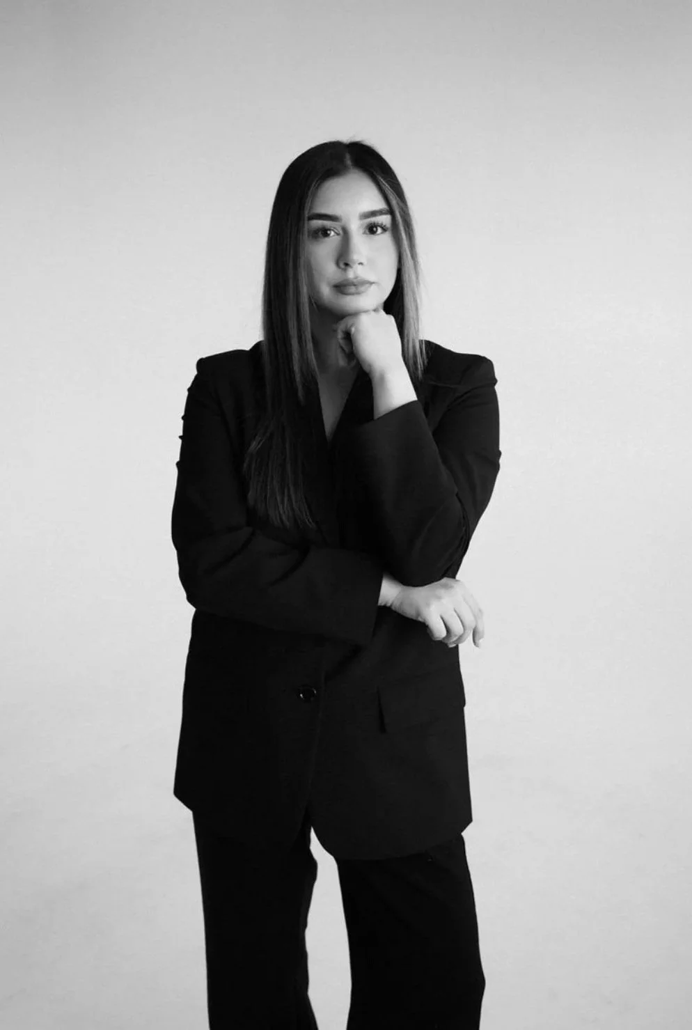 Black and white portrait of a woman with long dark hair, wearing a blazer, standing with her chin resting on her hand against a plain background.