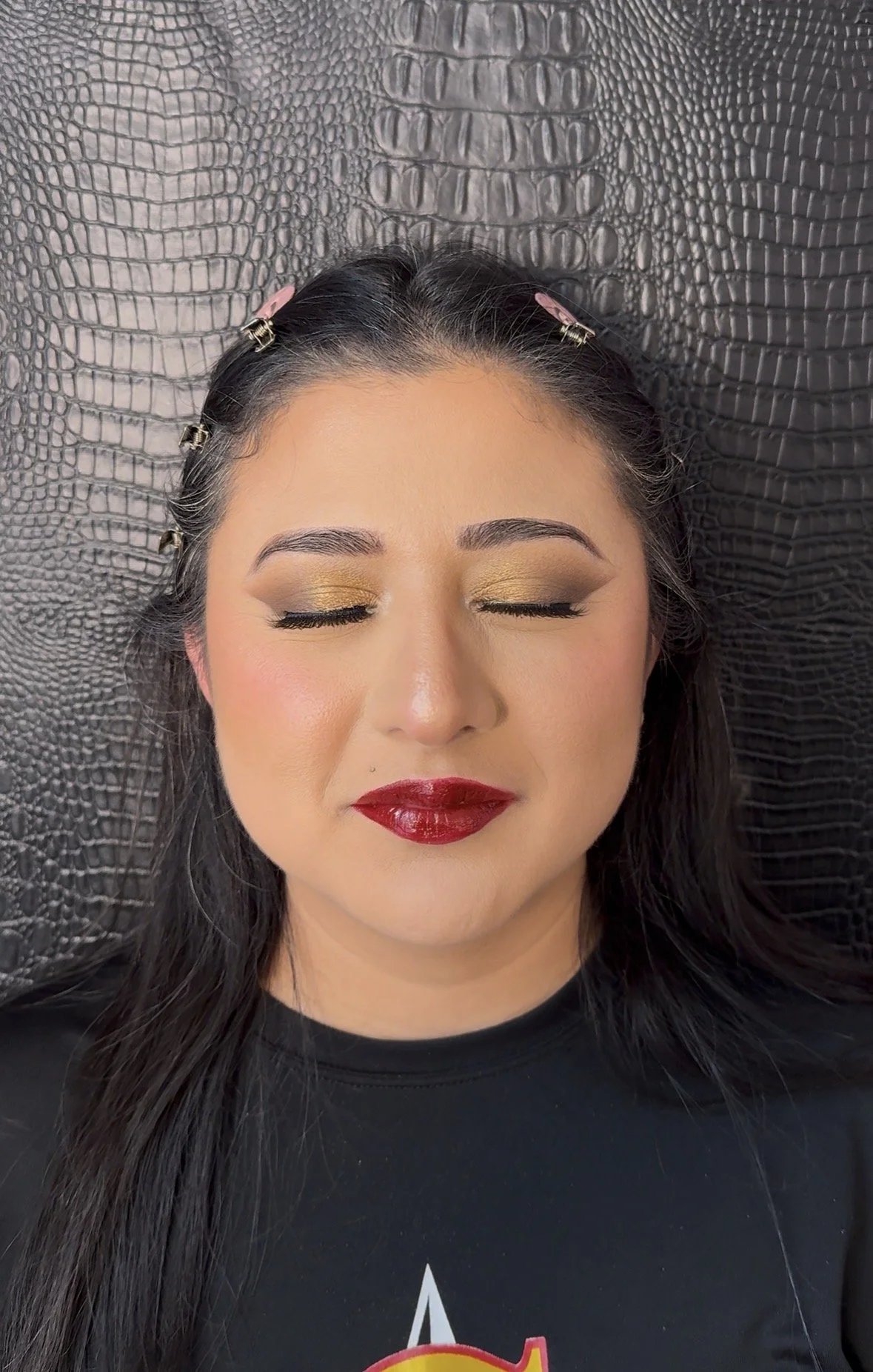 A woman with closed eyes lying on a textured black surface, wearing gold eyeshadow, bold red lipstick, and hair clip pins.