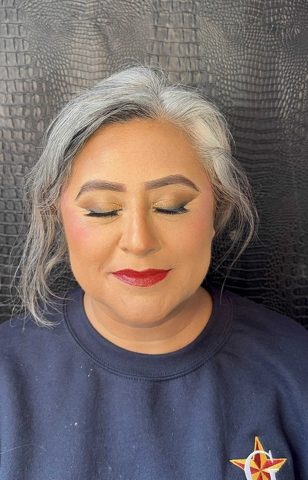 A woman with gray hair and closed eyes, wearing makeup with gold eyeshadow, eyeliner, and red lipstick, dressed in a dark sweatshirt with a NASA logo.
