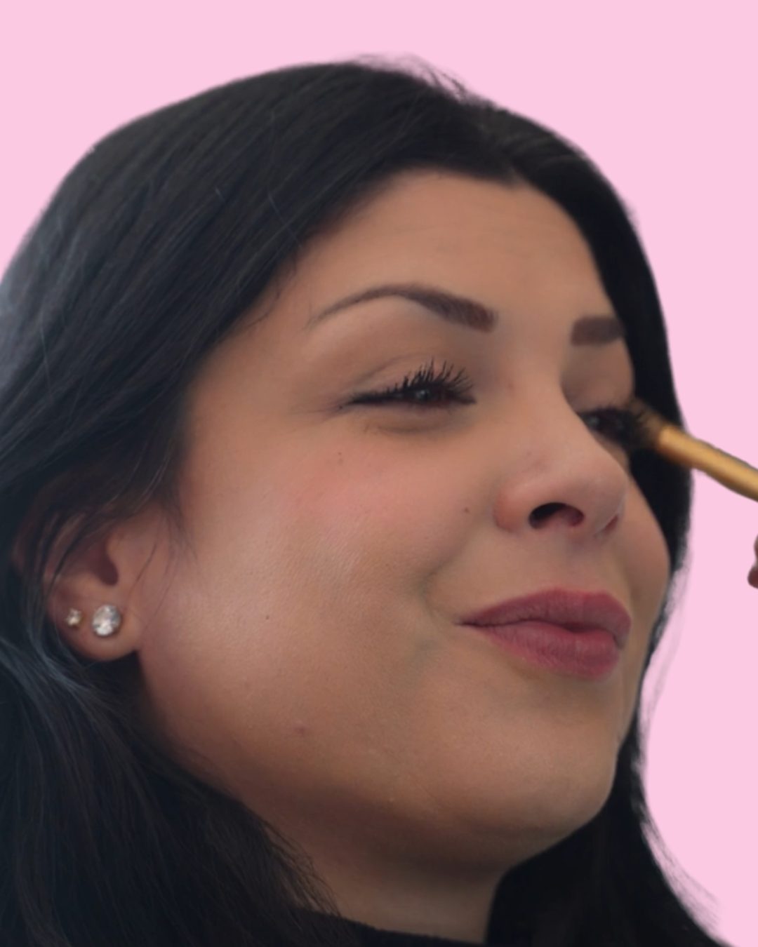 Close-up of a woman with dark hair getting eye makeup applied with a brush, pink background.