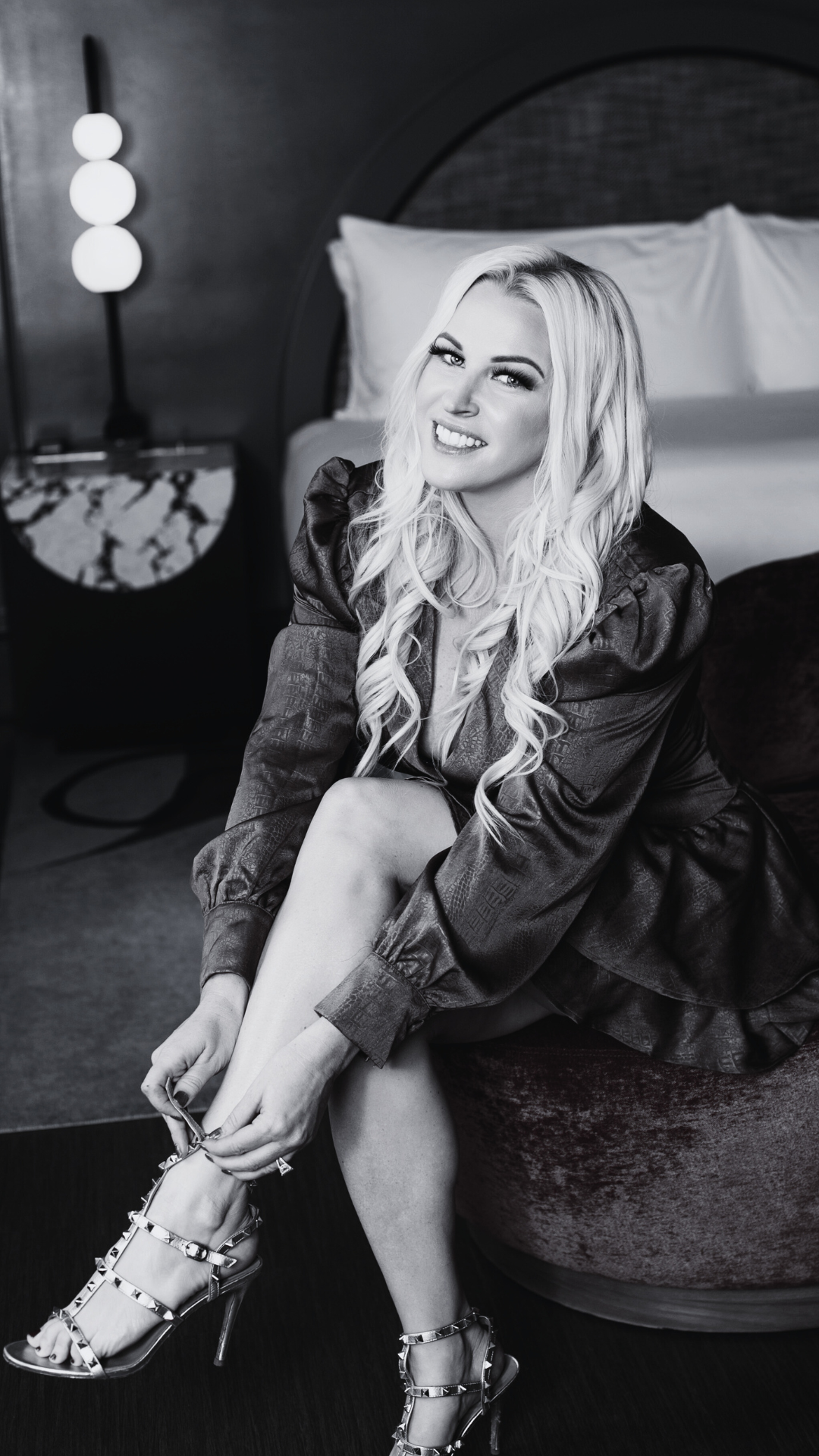 Black and white photo of a blonde woman sitting on a round ottoman, smiling and fixing her high-heeled shoes, in a bedroom setting.