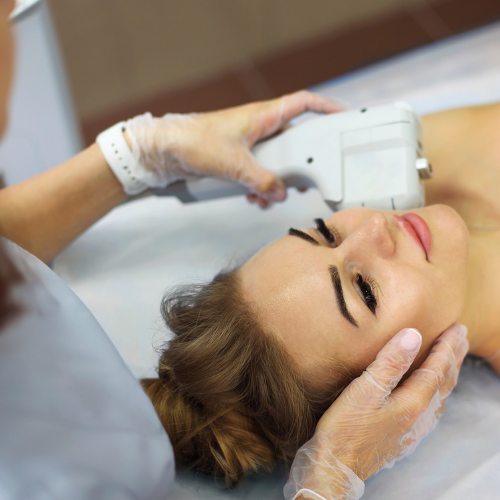 A woman receiving a non-invasive cosmetic treatment using a handheld device on her forehead, while lying on a treatment bed with a medical professional holding the device and supporting her head.