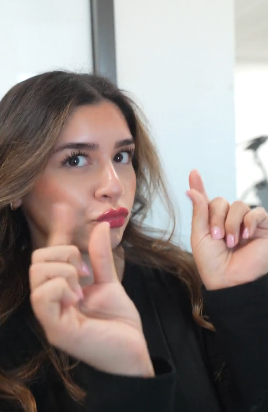 A woman with long, wavy brown hair and makeup, posing with her fingers in a pointing gesture near her face, indoors with a plain background.