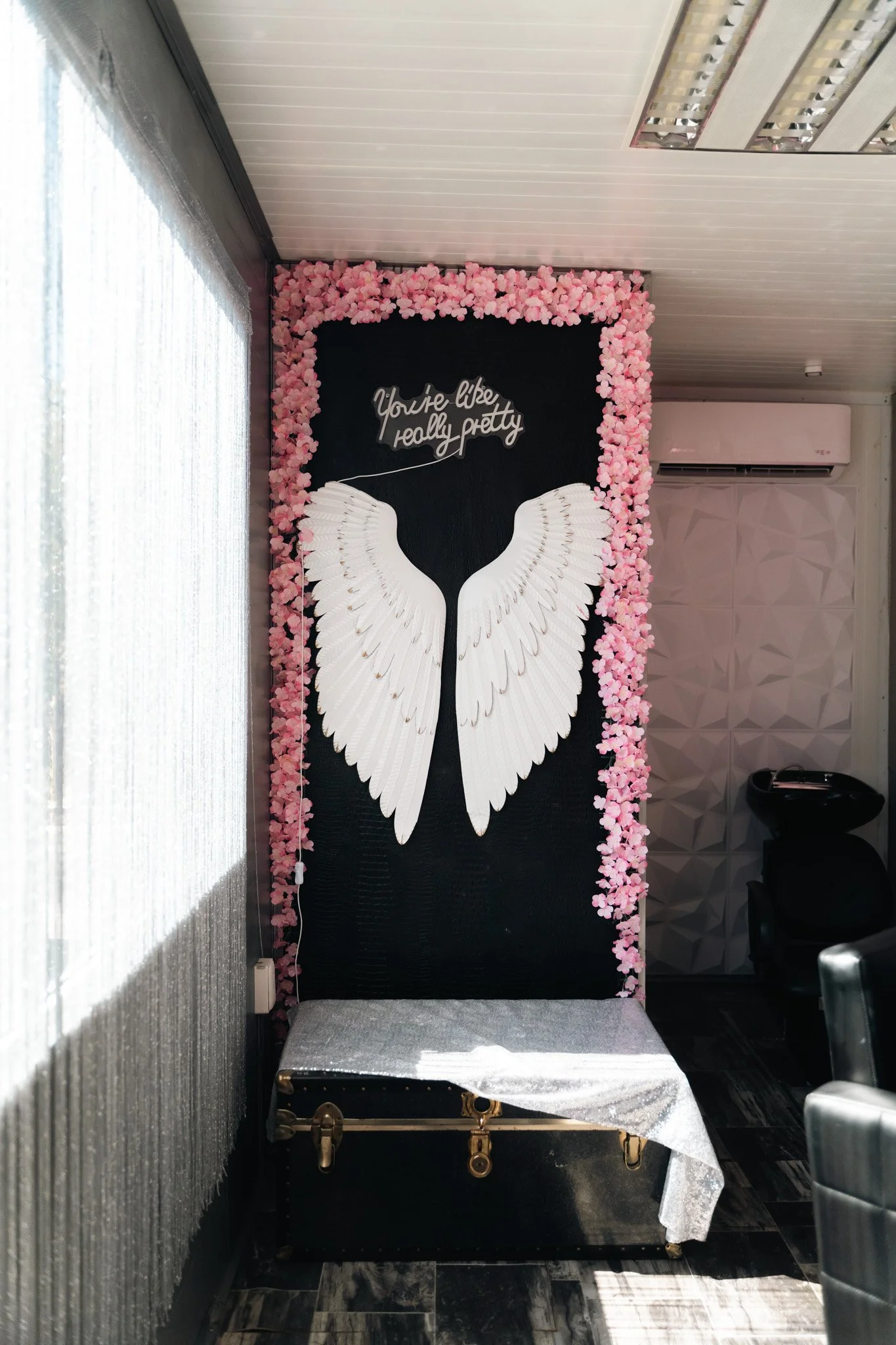 Decorative wall with pink flowers, white angel wings, and a sign that says 'You're like really pretty'; includes a black trunk covered with a cloth.