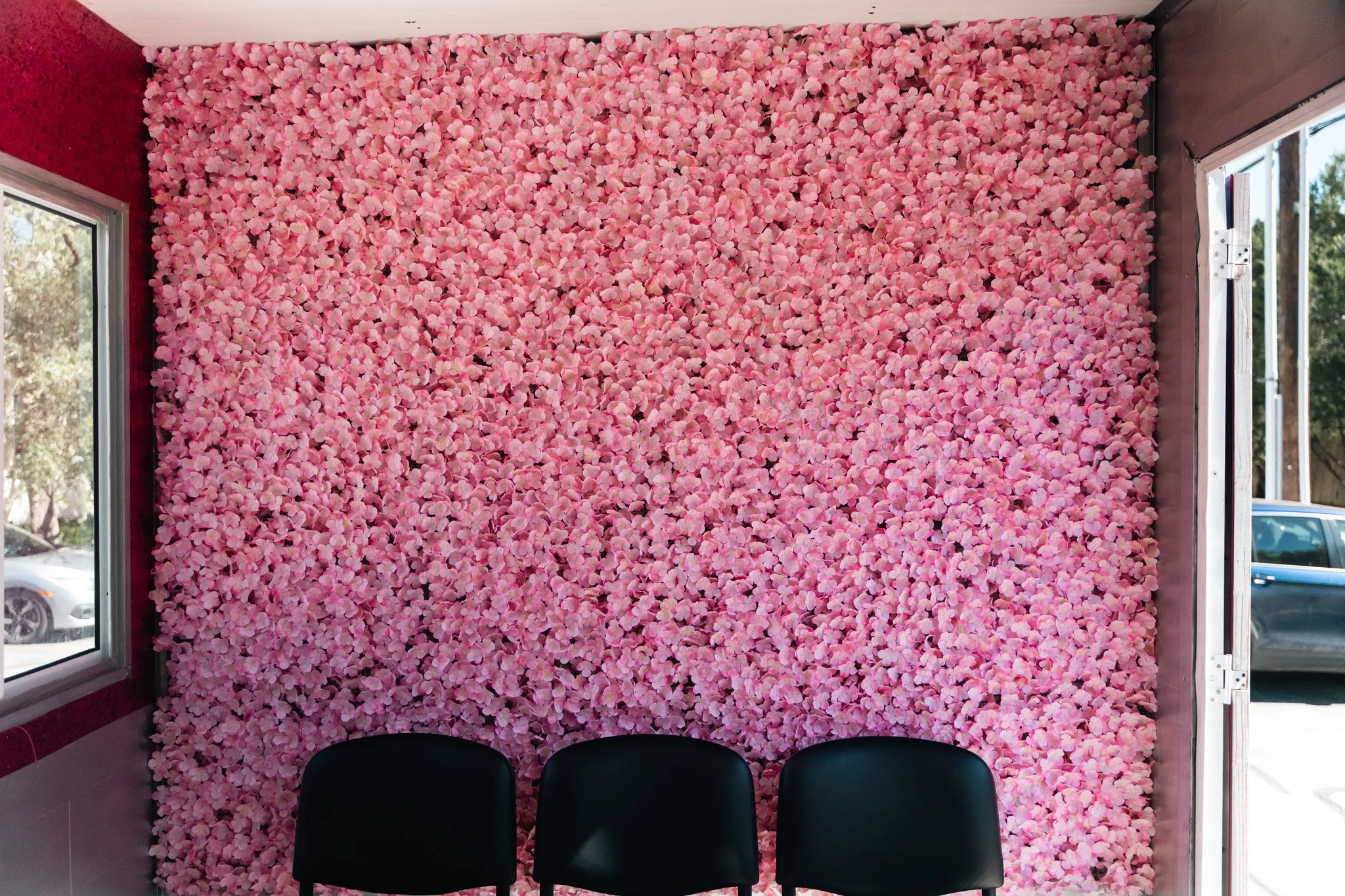 Pink floral wall with three black chairs in front of it, window on the left, and open door on the right showing cars outside.