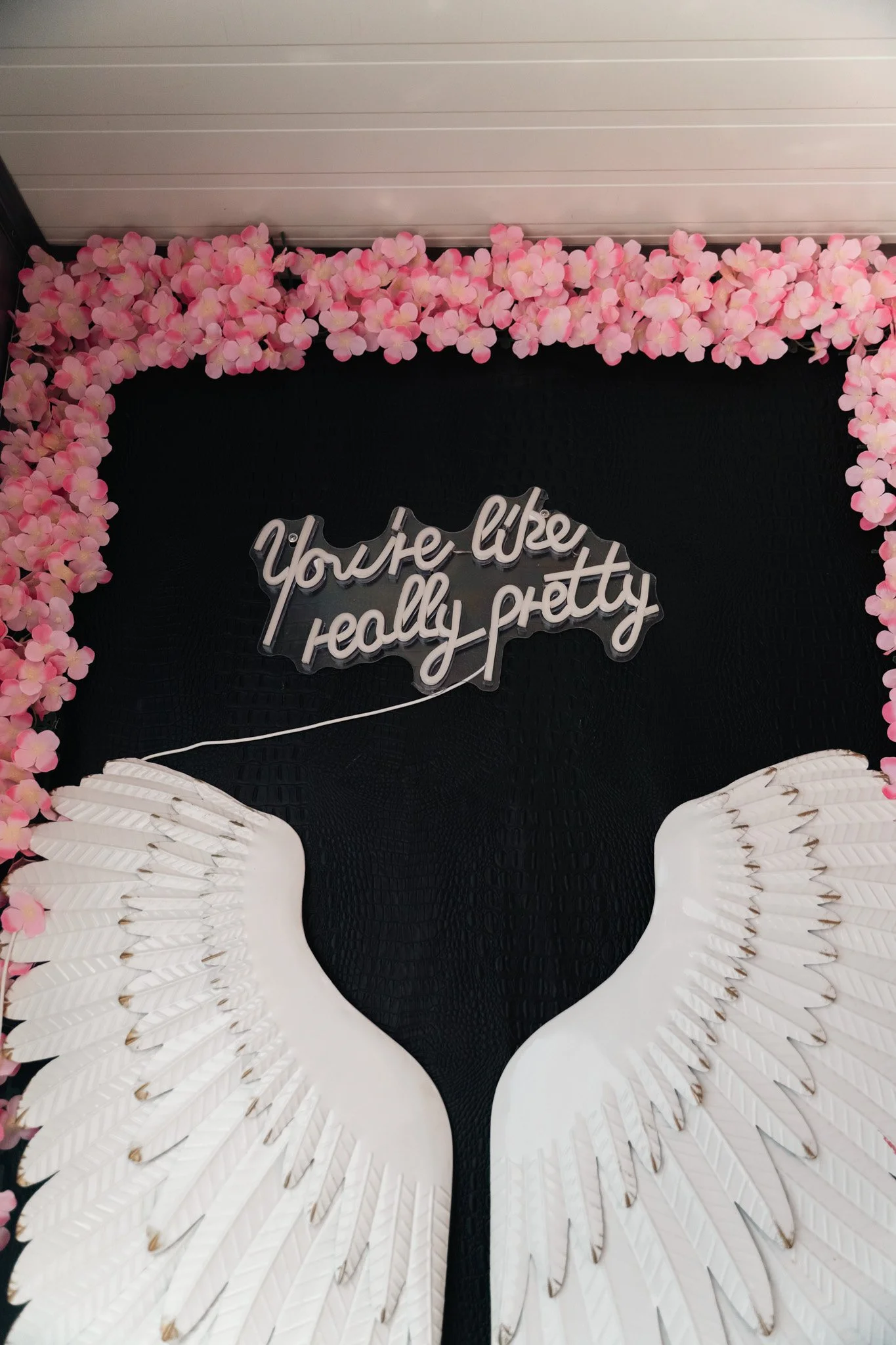 Pink flower garland framing a sign that reads 'You're like really pretty' with white decorative wings below.