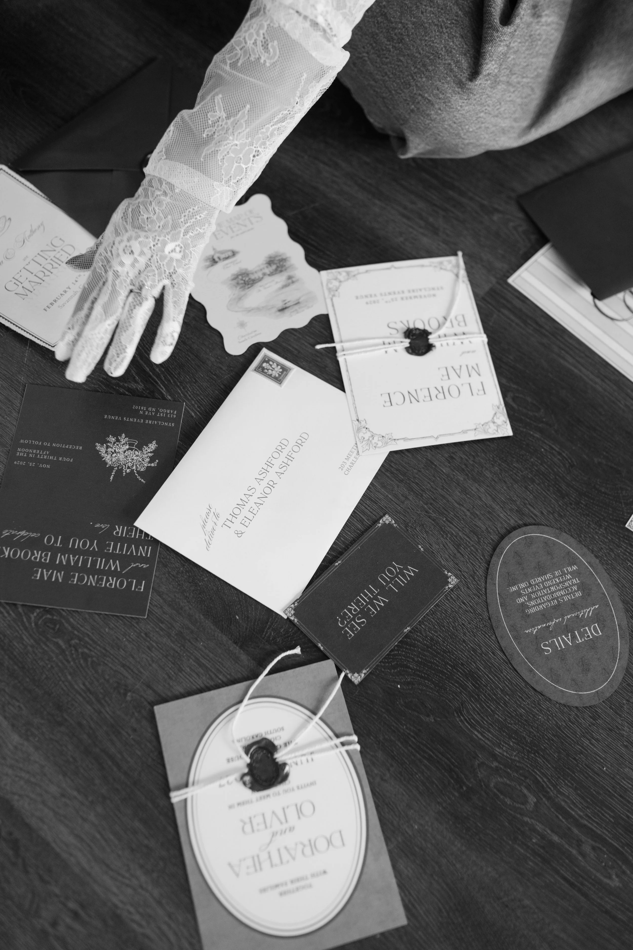 Black and white photo of wedding and engagement invitation cards laid out on a wooden table, with one card featuring a lace-gloved hand pointing towards the cards.