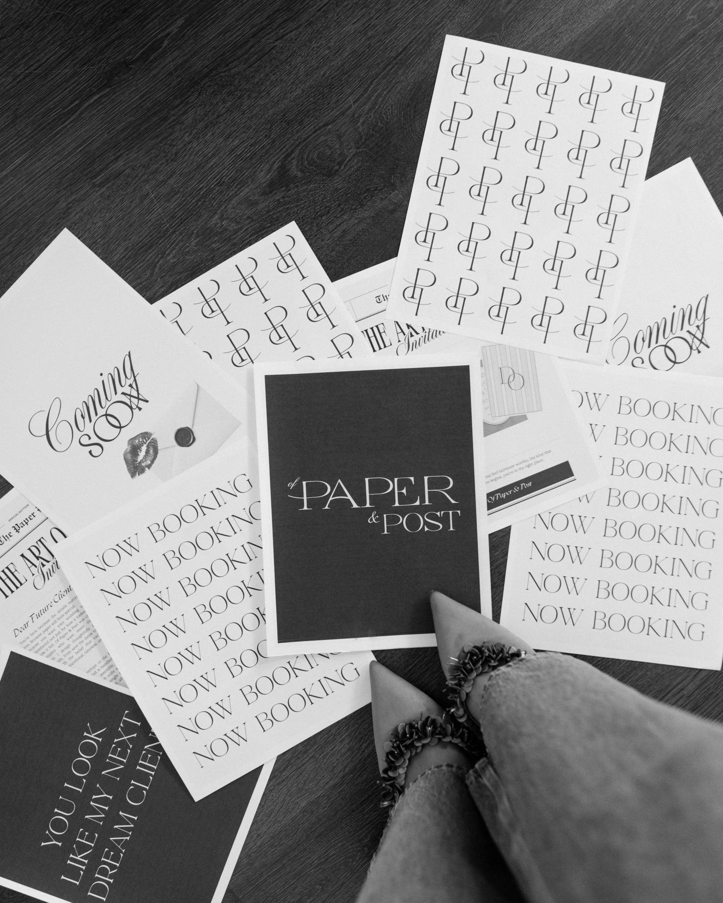 A person's hand with a decorated bracelet reaching towards promotional flyers on a dark wooden surface. The flyers include text about booking and a paper and post themed event.