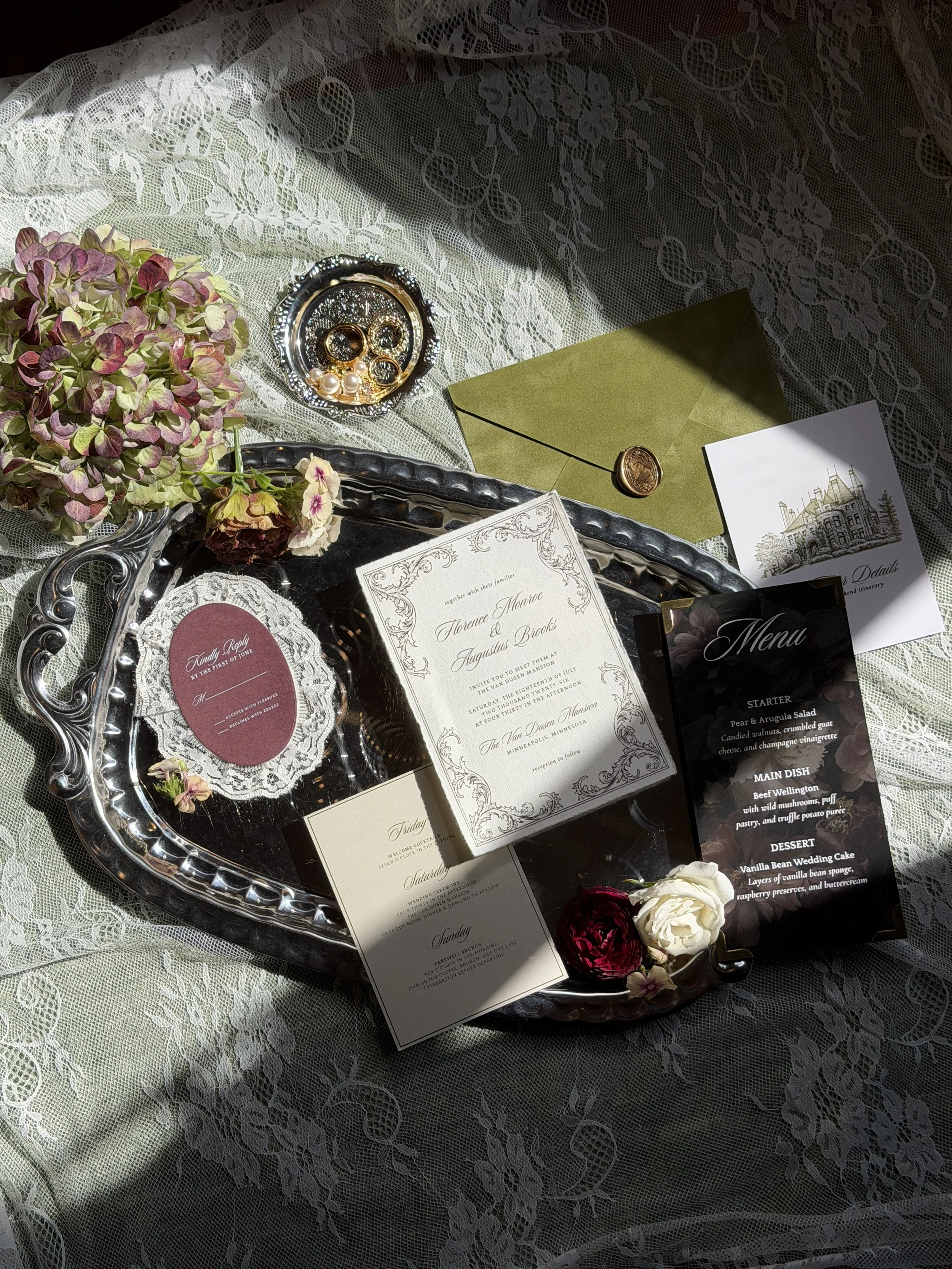 A wedding reception setup featuring a silver tray with flowers, rings in a small dish, and an olive green envelope, all placed on a lace tablecloth with wedding invitations, a menu card, and decorative flowers.