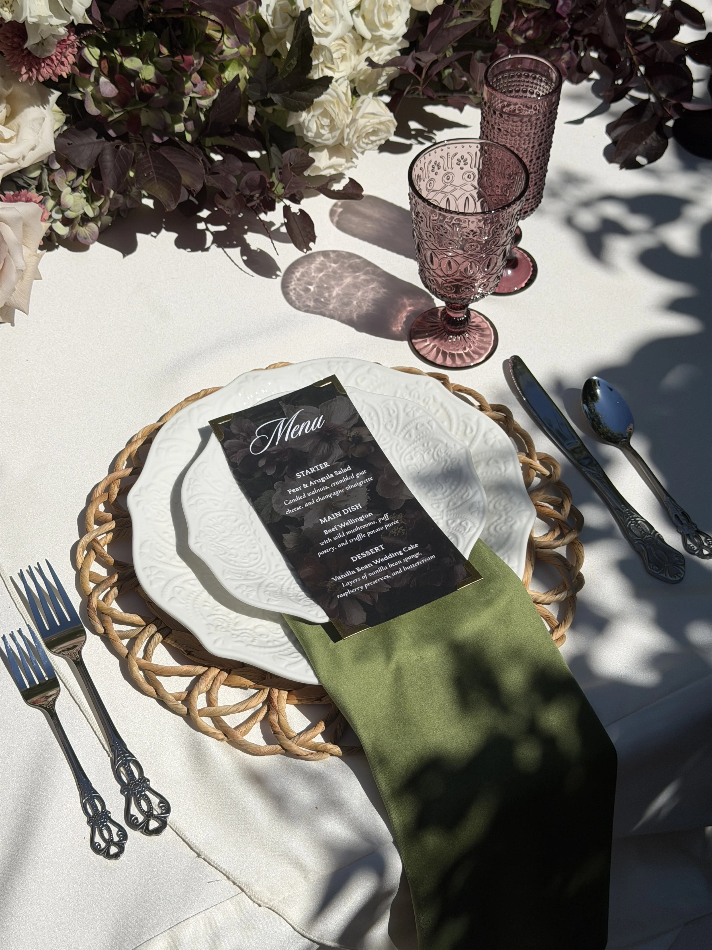 A table setting with a white plate, silver cutlery, two pink textured glasses, a menu, a green napkin, and decorative flowers in the background.