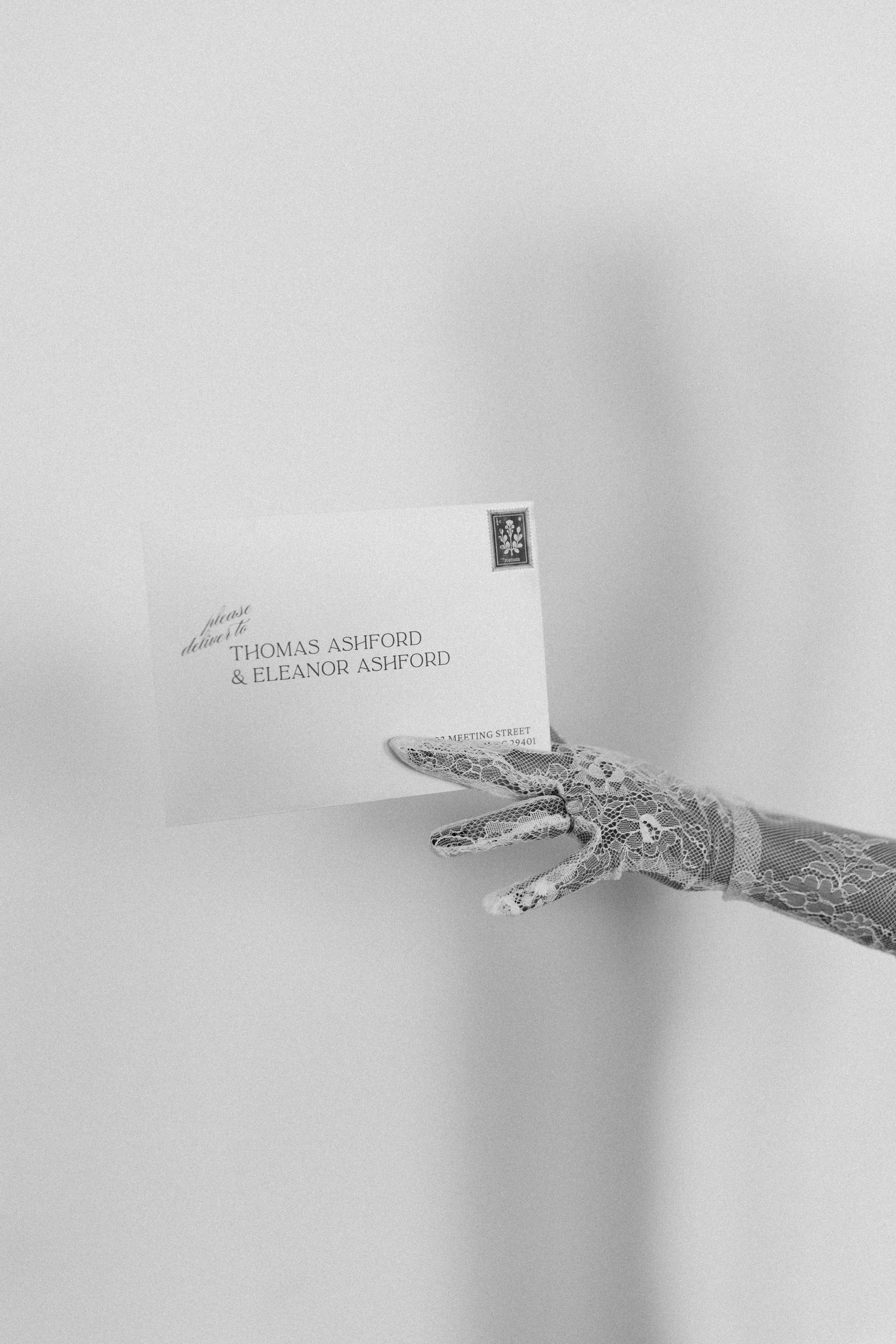 Wedding invitation card held by a lace-gloved hand, on a plain background.