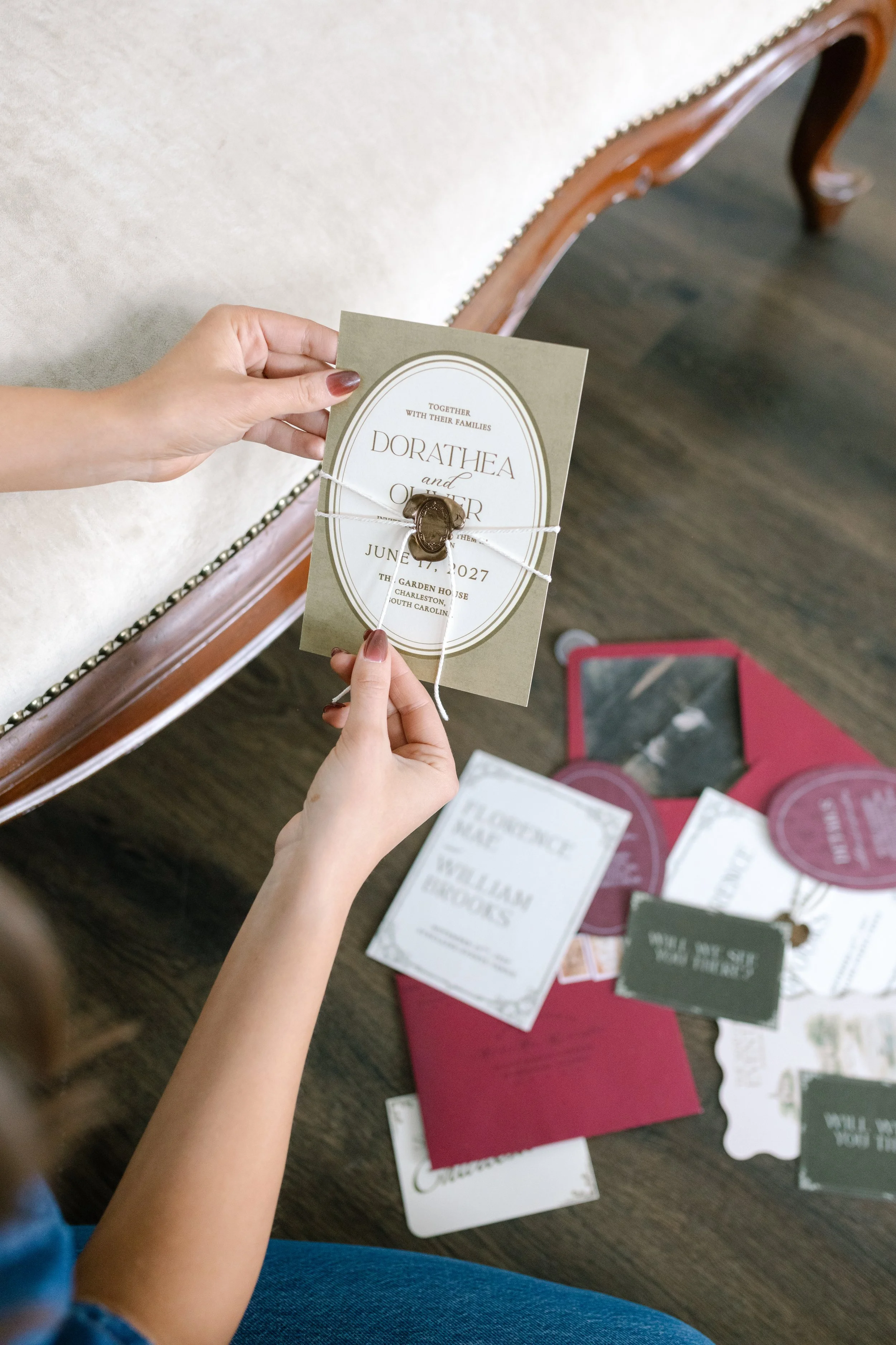 A person holding a wedding invitation with a wax seal and string, surrounded by various stationery on a wooden table.