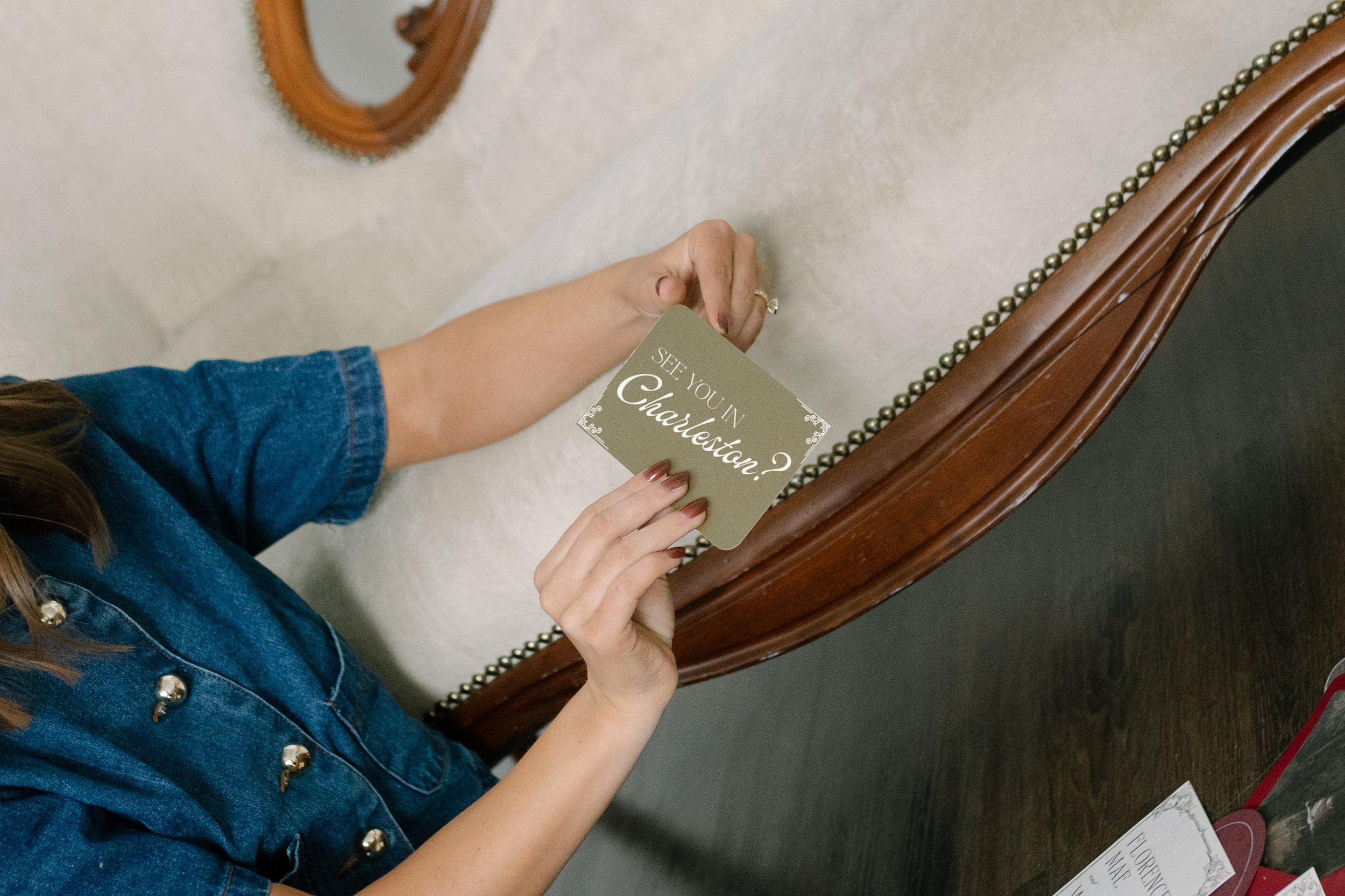 A woman wearing a denim jacket holding a card that says, 'See you in Charlestown?' while seated at a wooden table with a mirror, papers, and a pen on it.