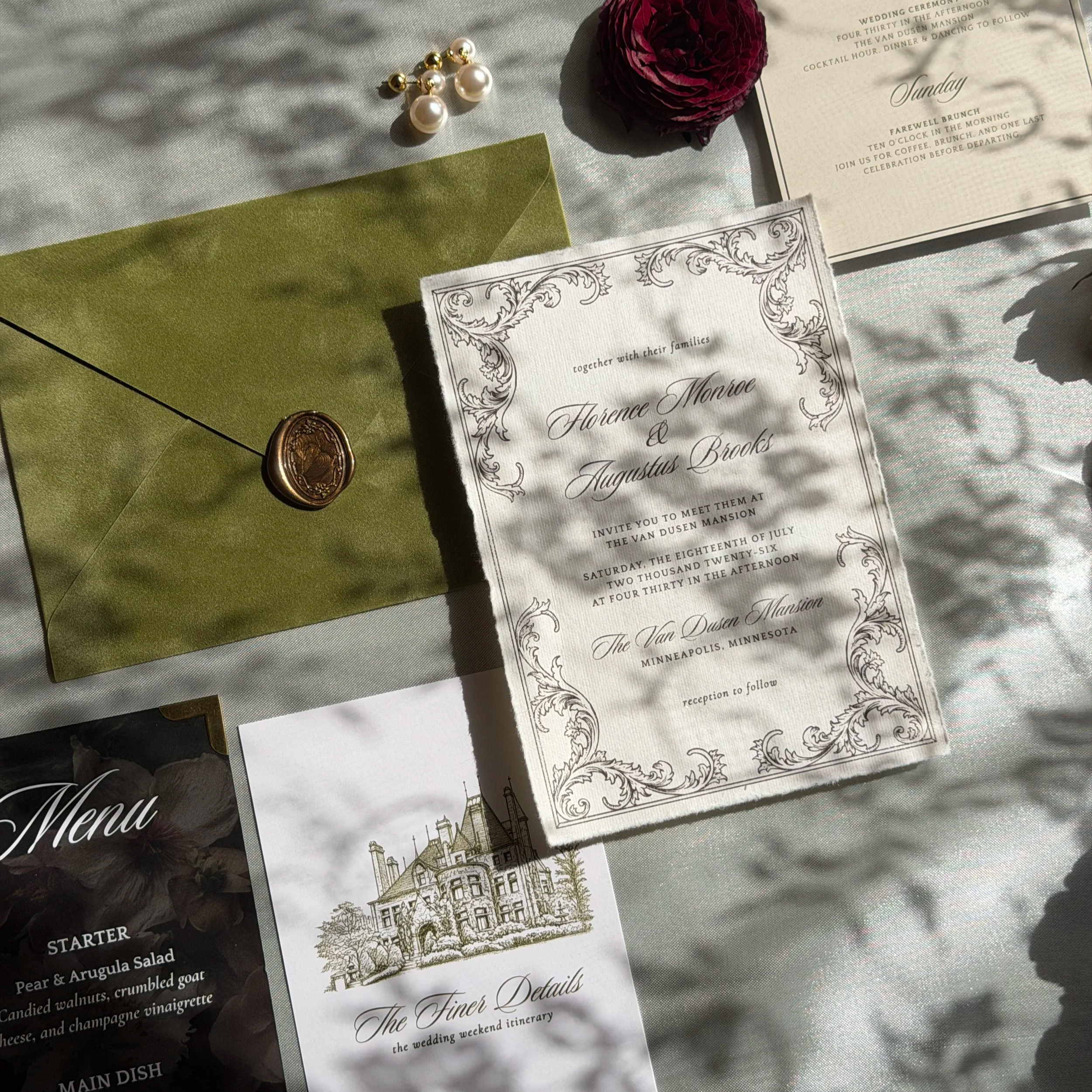 A wedding menu with a green envelope, a pair of pearl and gold earrings, a dark red rose, and an illustrated wedding invitation featuring a house and decorative border, all placed on a satin-like fabric.