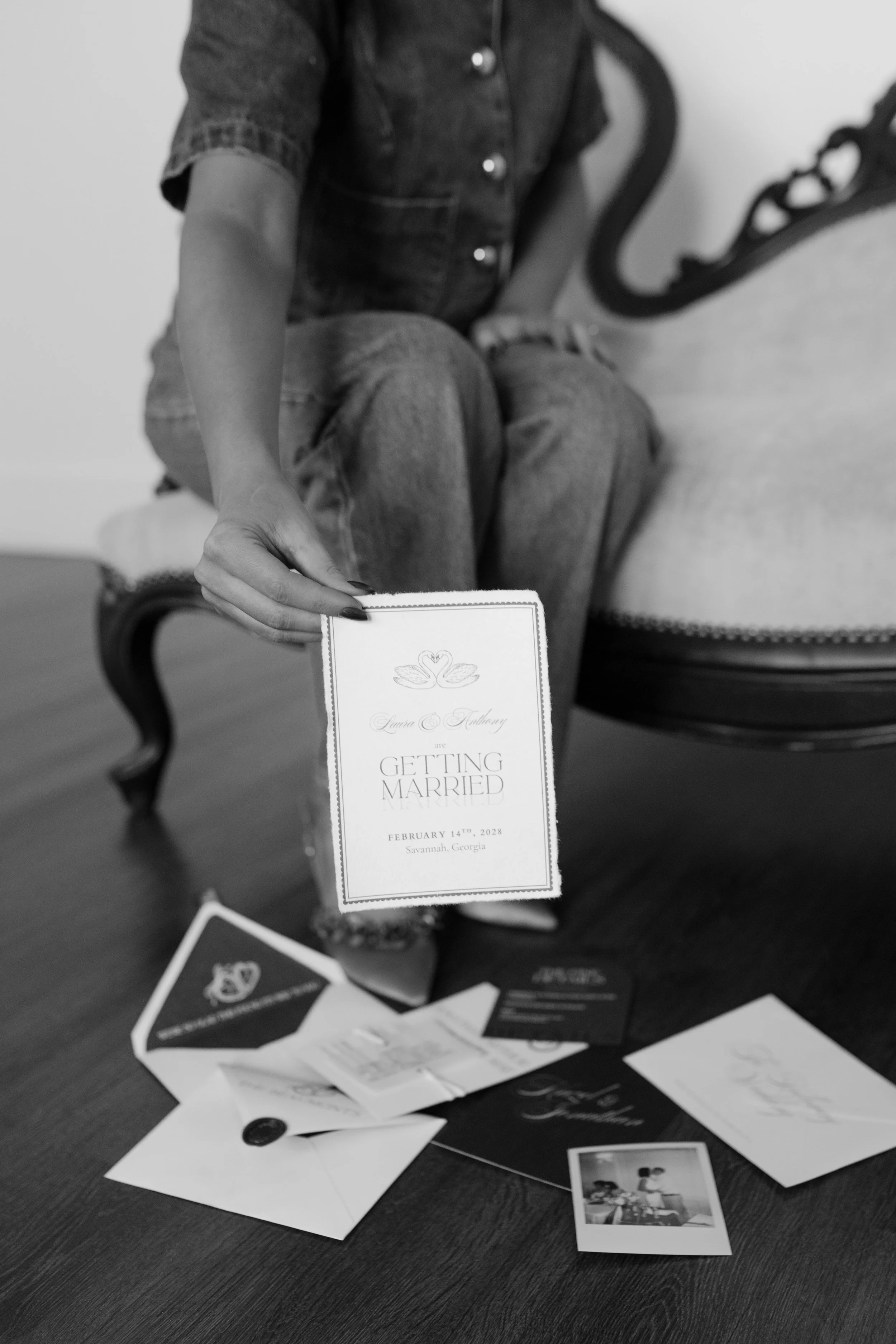 A woman sitting on a vintage chair displays a wedding invitation with wedding-related cards and a photo on the table below.