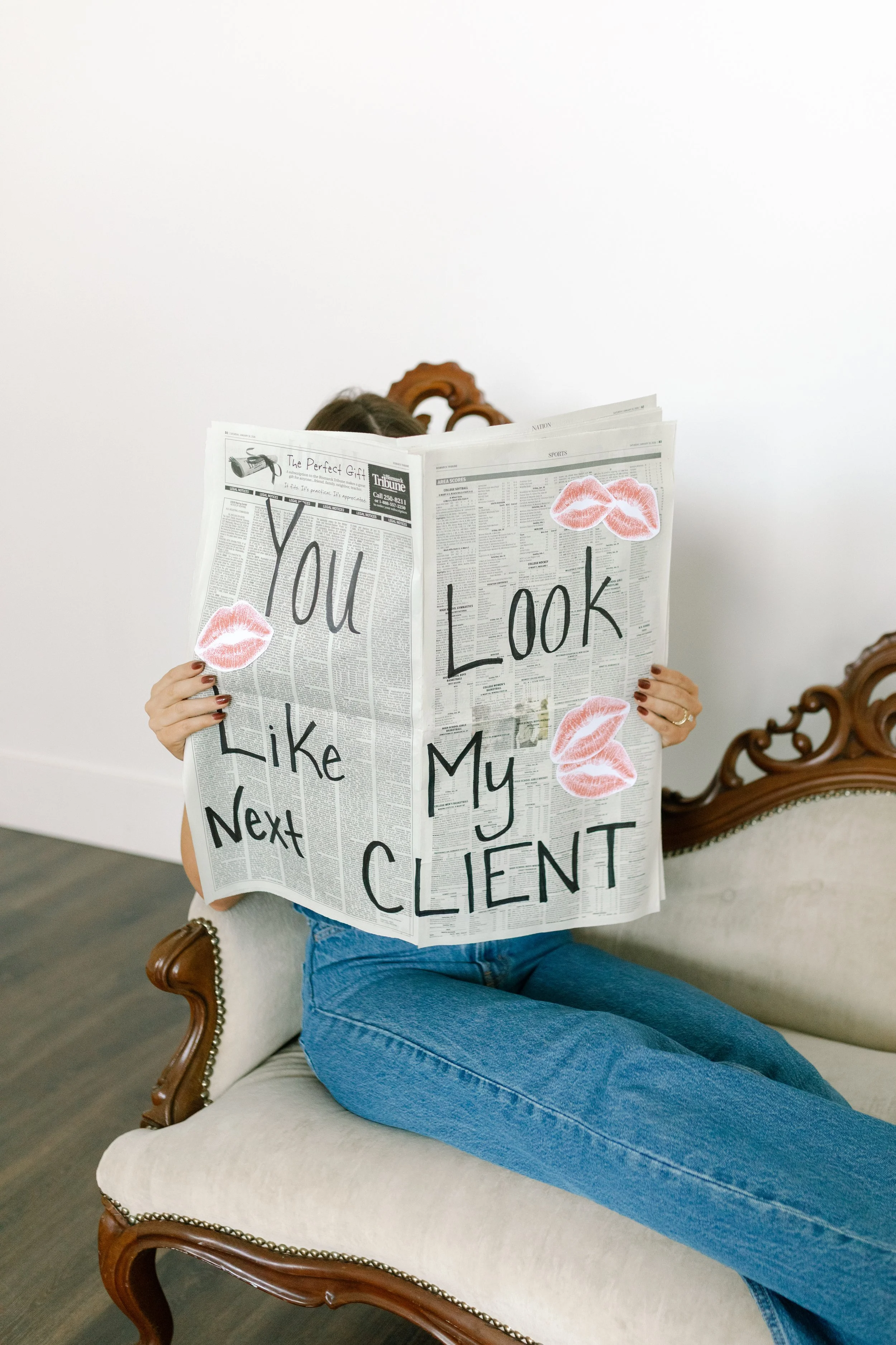 Person sitting on a beige upholstered vintage-style sofa holding a newspaper that reads 'You Look Like My Next Client'. The person is wearing blue jeans, and their face is hidden behind the newspaper decorated with pink lipstick kiss stickers. The sofa has a wooden carved frame and a white wall is in the background.