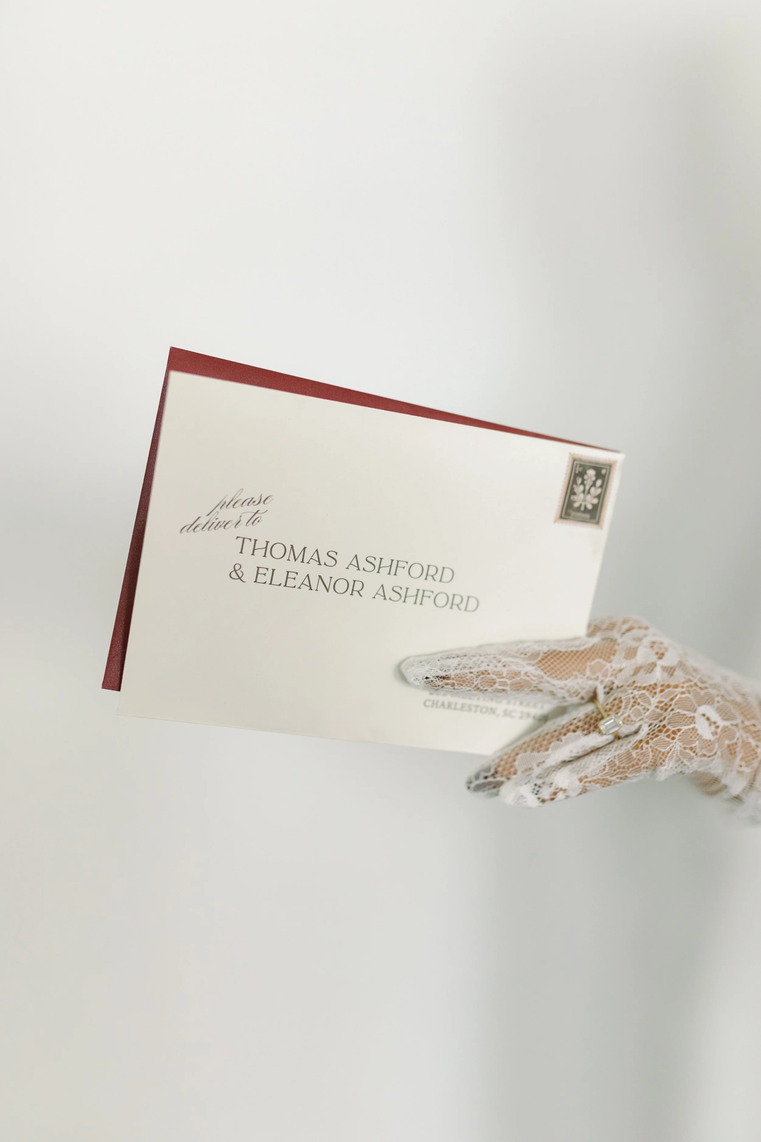 A wedding invitation card held by a lace-gloved hand, with a gray and white floral design at the top right corner and elegant script text reading 'please deliver to Thomas Ashford & Eleanor Ashford'.