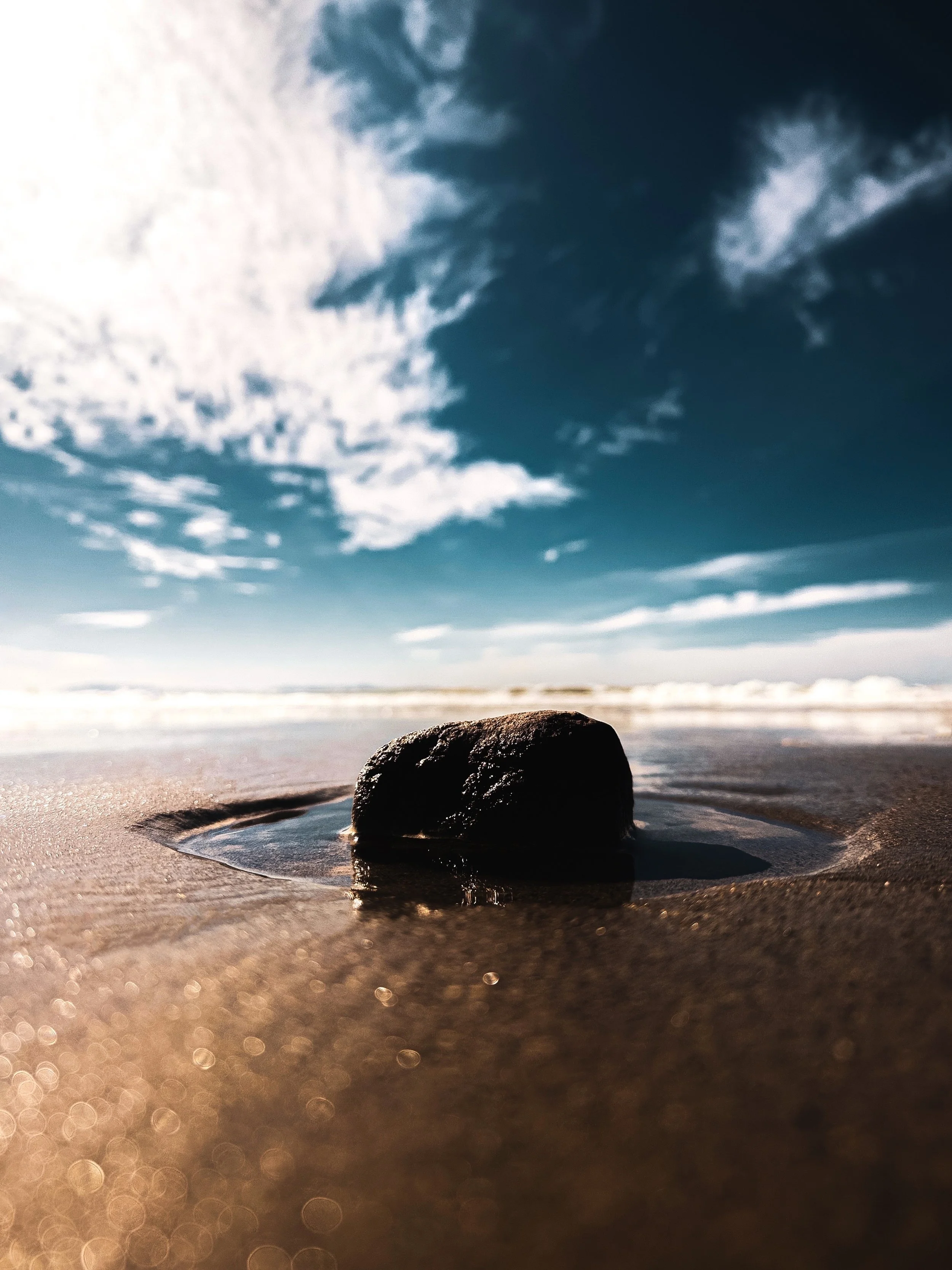 A close-up view of a black rock partially submerged in wet sand on a beach, with a cloudy sky overhead and a horizon of ocean waves in the distance.
