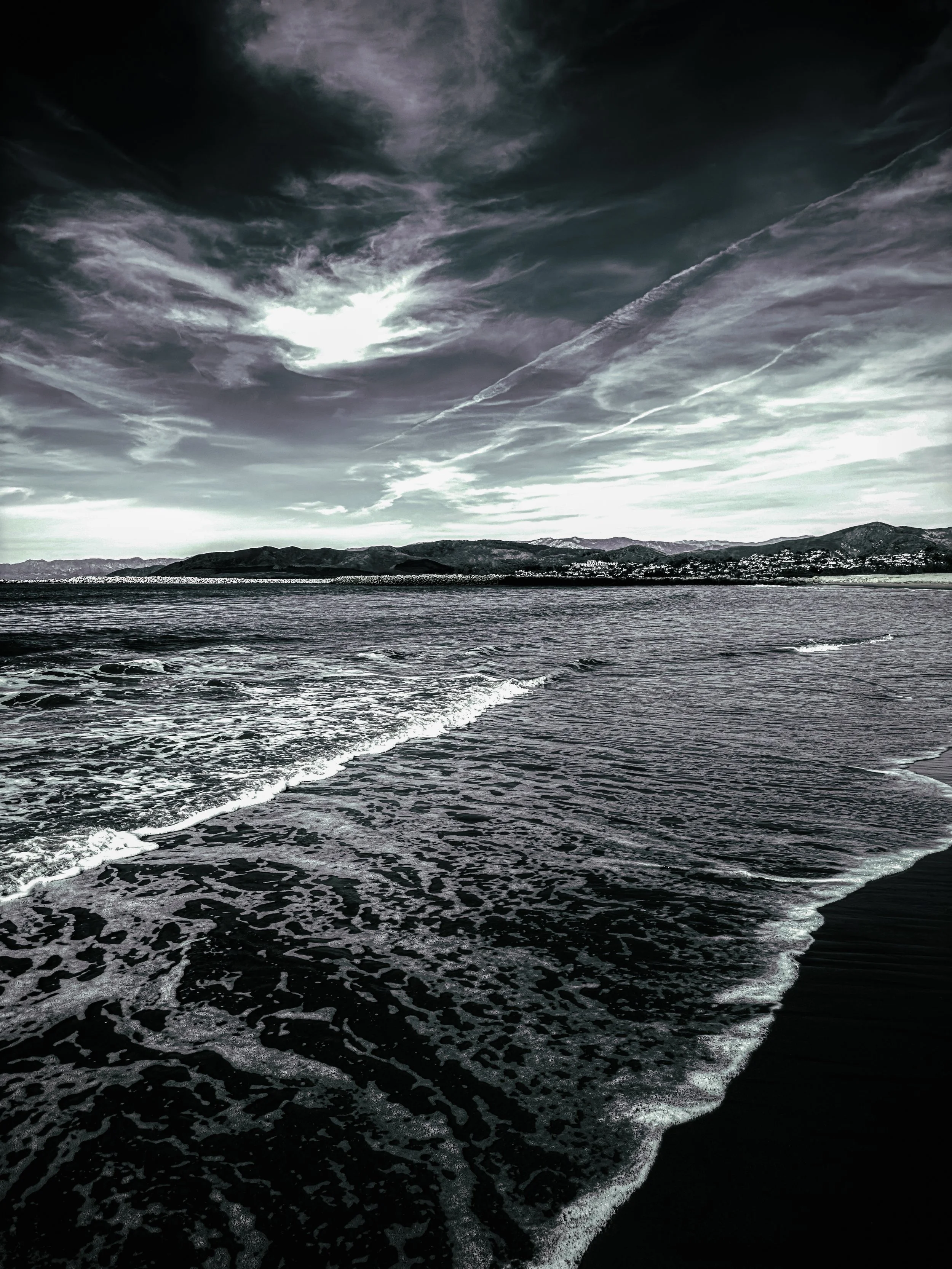 A black and white photograph of a beach scene with waves washing onto the shore and a cloudy sky above.