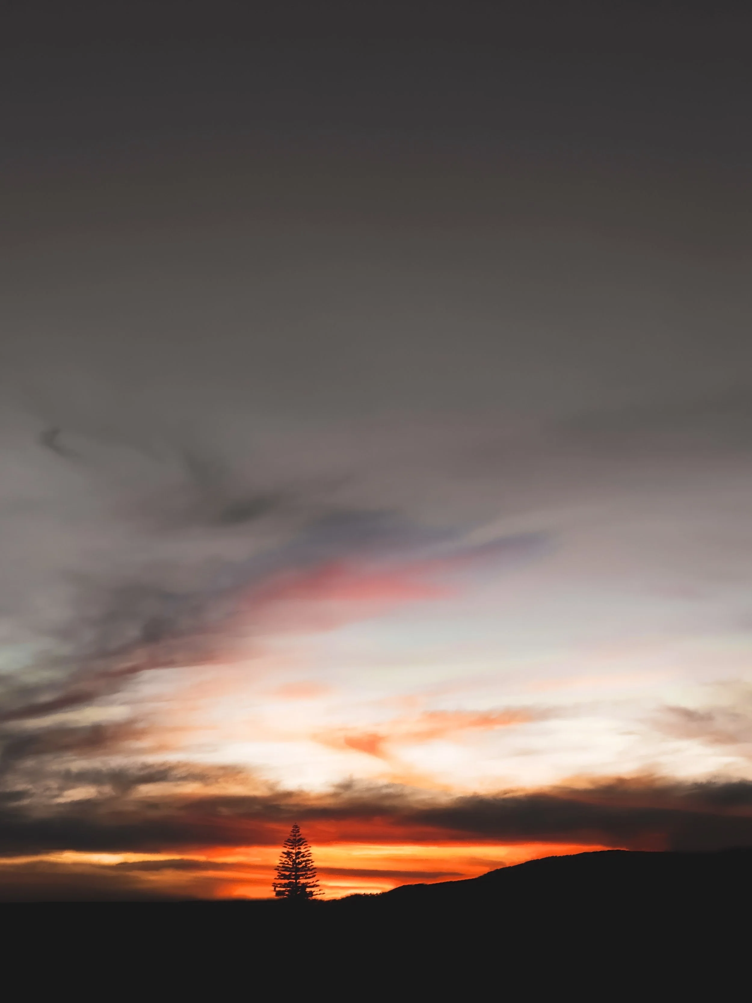 Sunset over a silhouette of a tree and hills with colorful clouds in the sky.