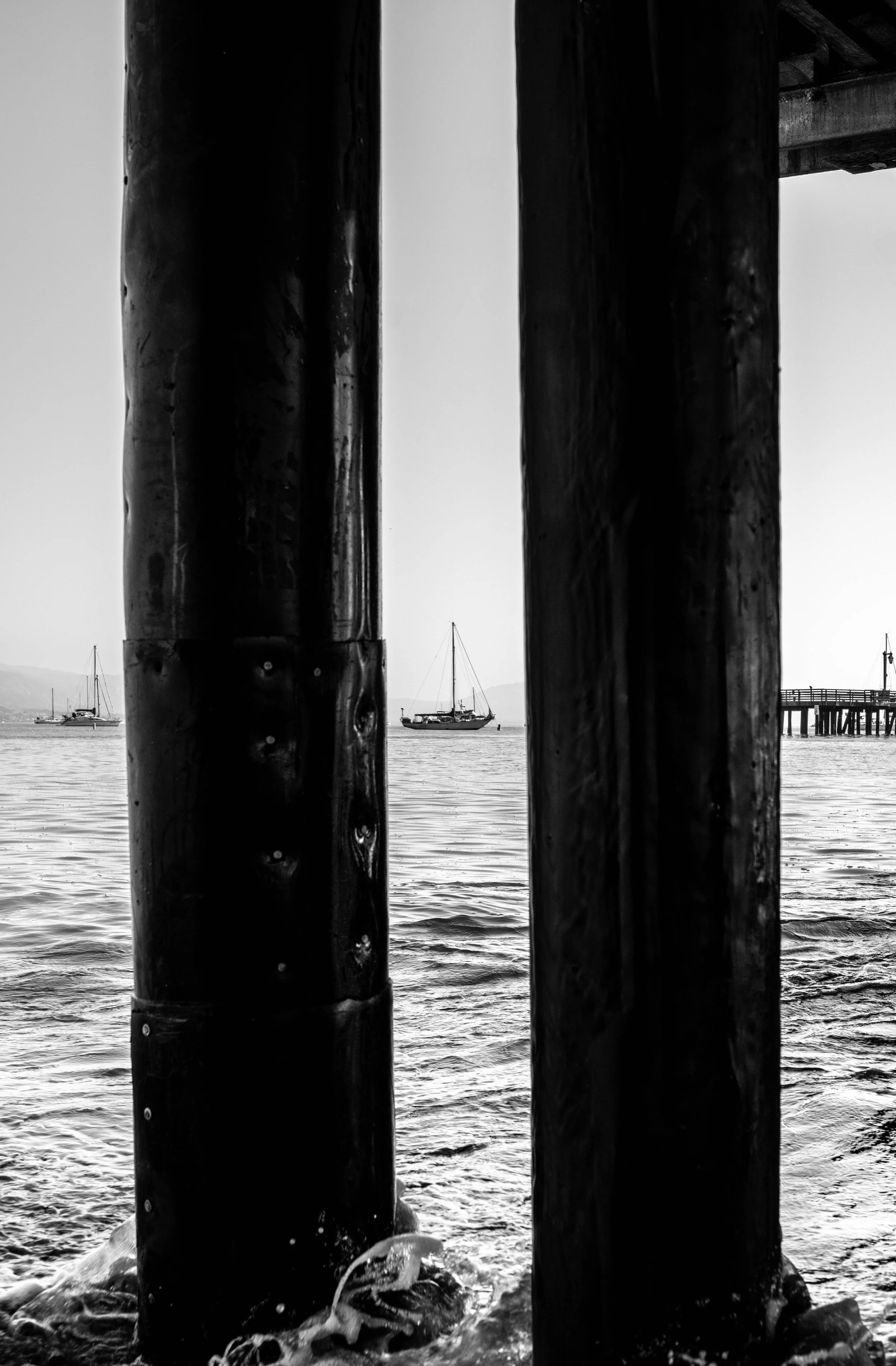 Black and white photo of a waterfront pier, with two large wooden pilings in the foreground and boats anchored in the water in the background.