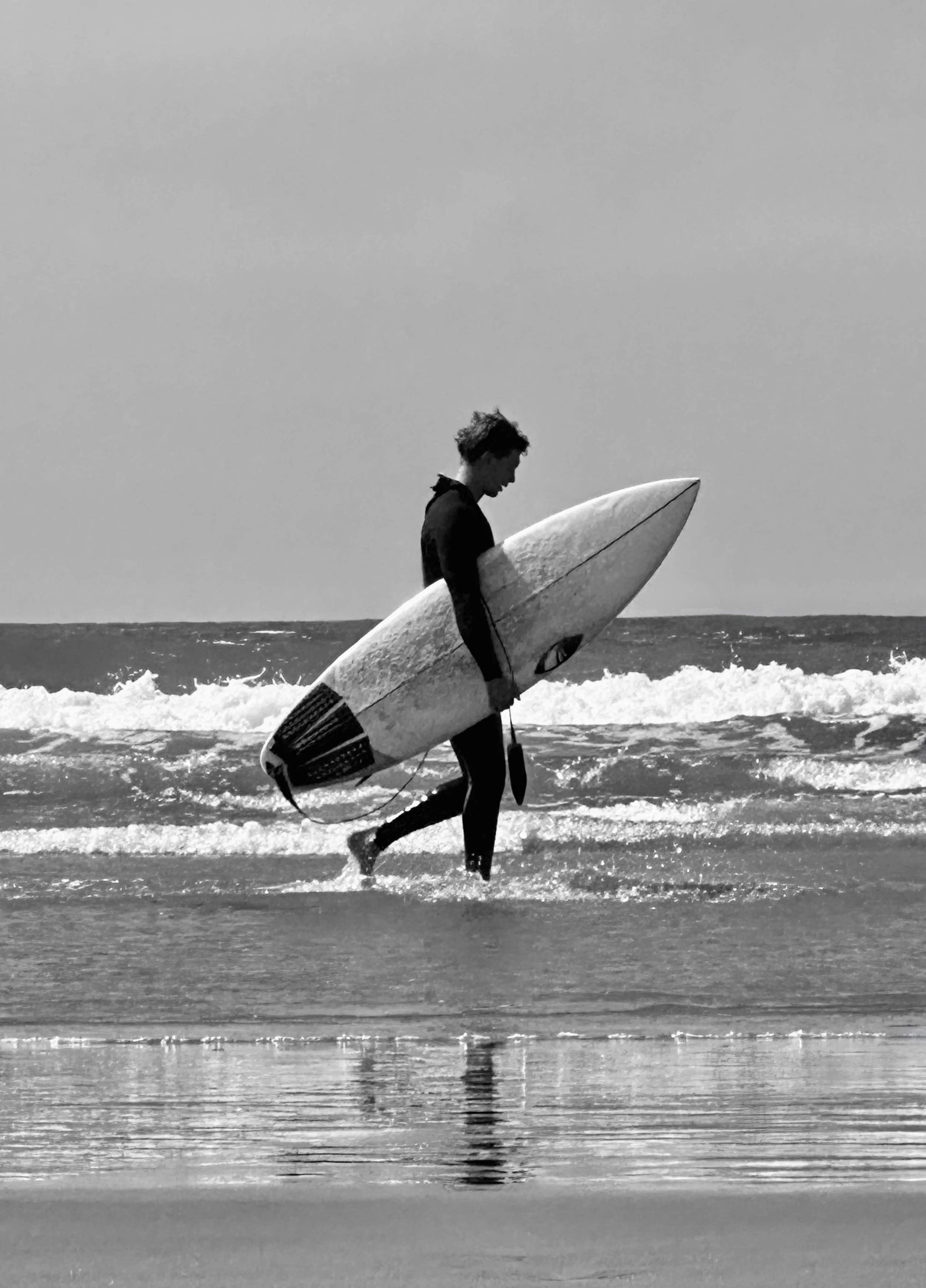 A person in a wetsuit walking in the shallow ocean water while holding a surfboard.