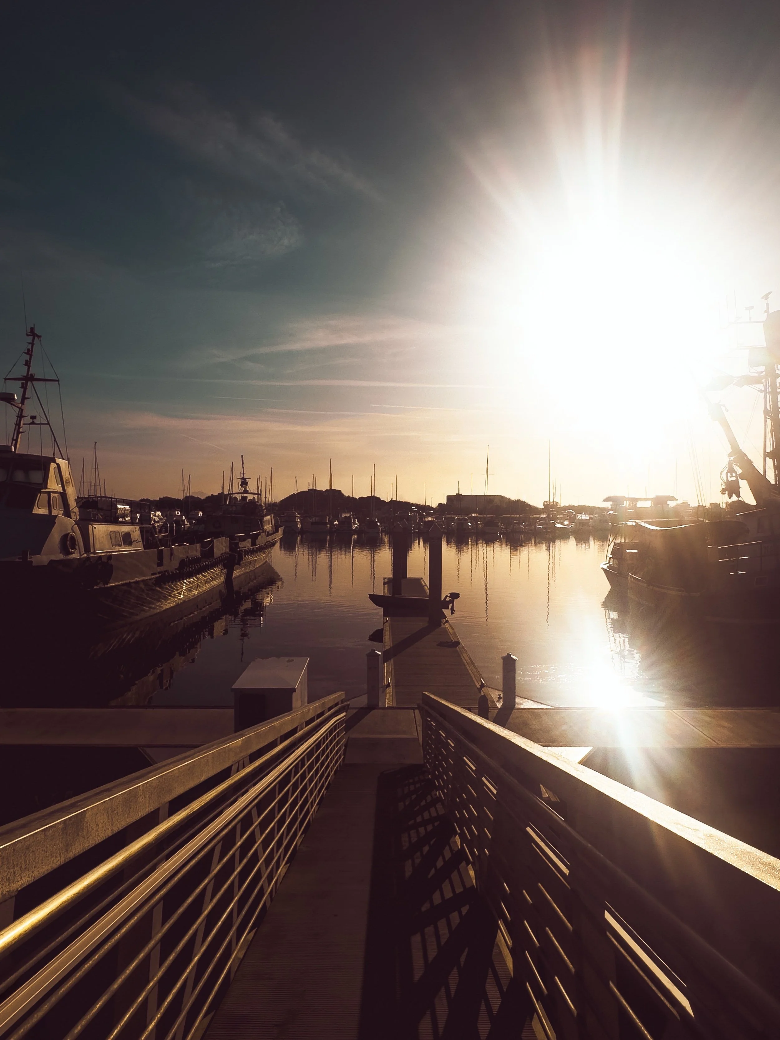 Marina with boats docked, a wooden walkway leading to the water, and the sun shining brightly in the sky creating lens flare.