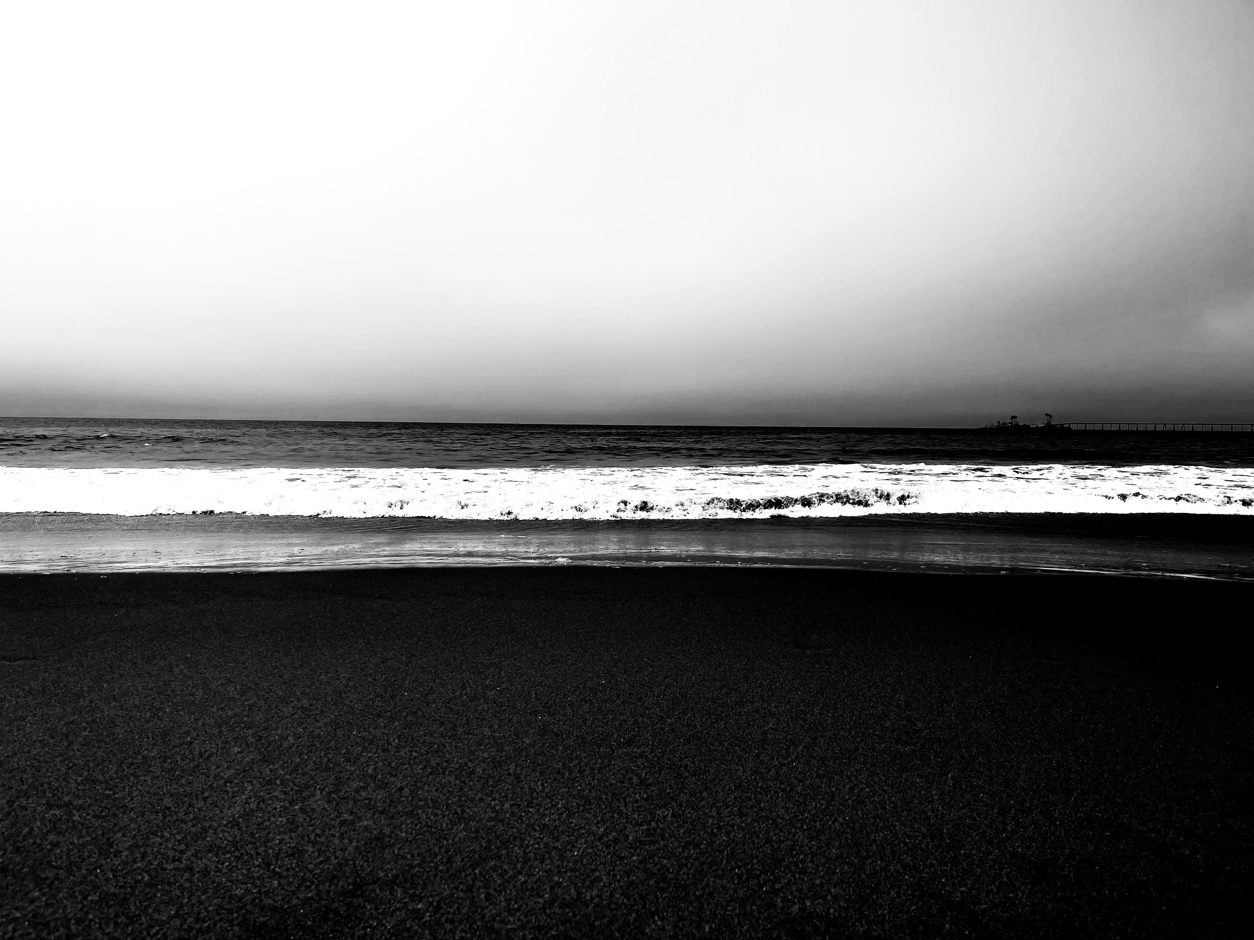Black and white photo of a beach with sand in the foreground, waves in the middle, and the ocean under a cloudy sky in the background.