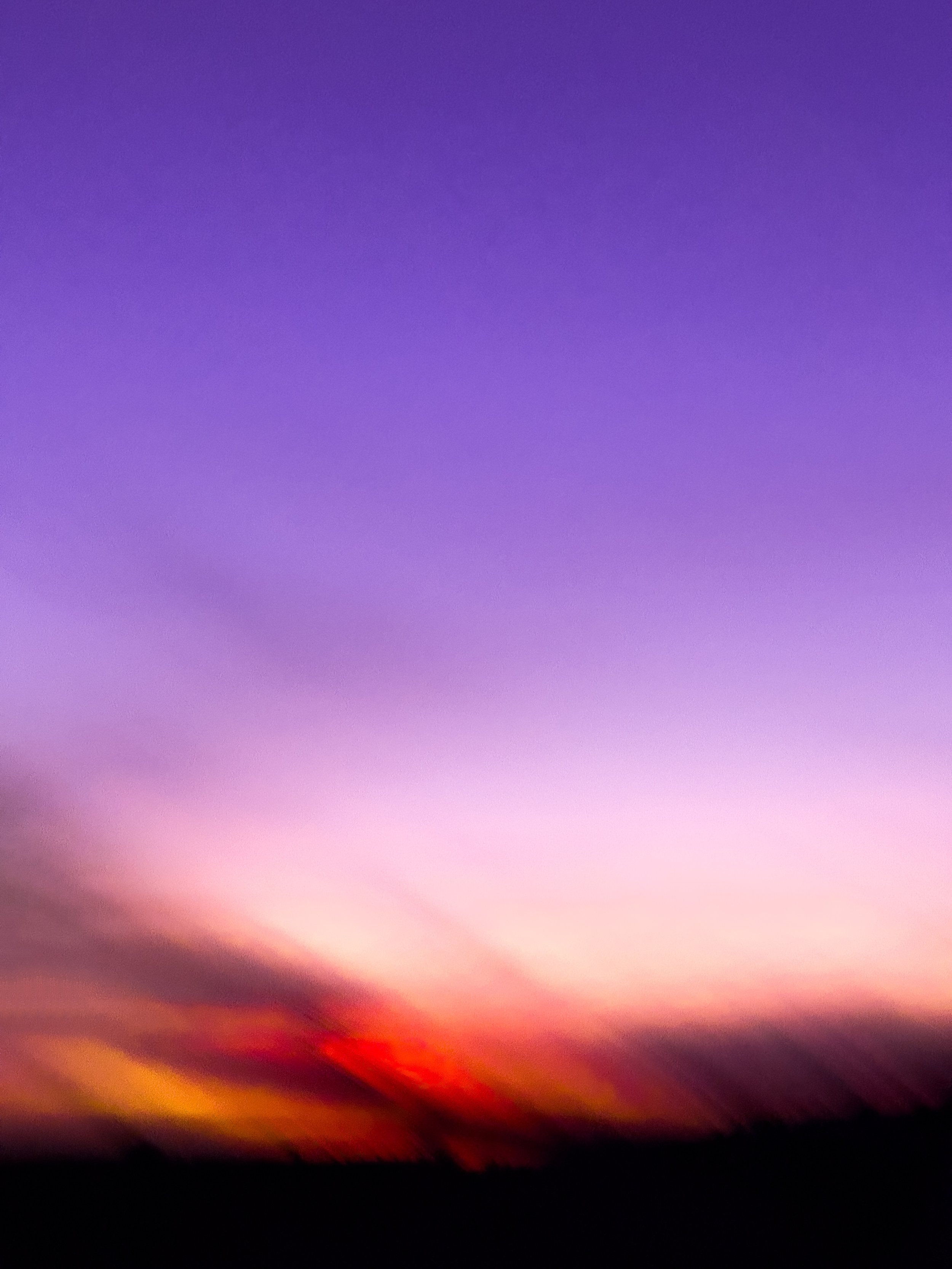 A colorful sunset or sunrise with a gradient sky transitioning from deep purple at the top to pink, orange, and red near the horizon, with dark silhouettes of trees or landscape at the bottom.