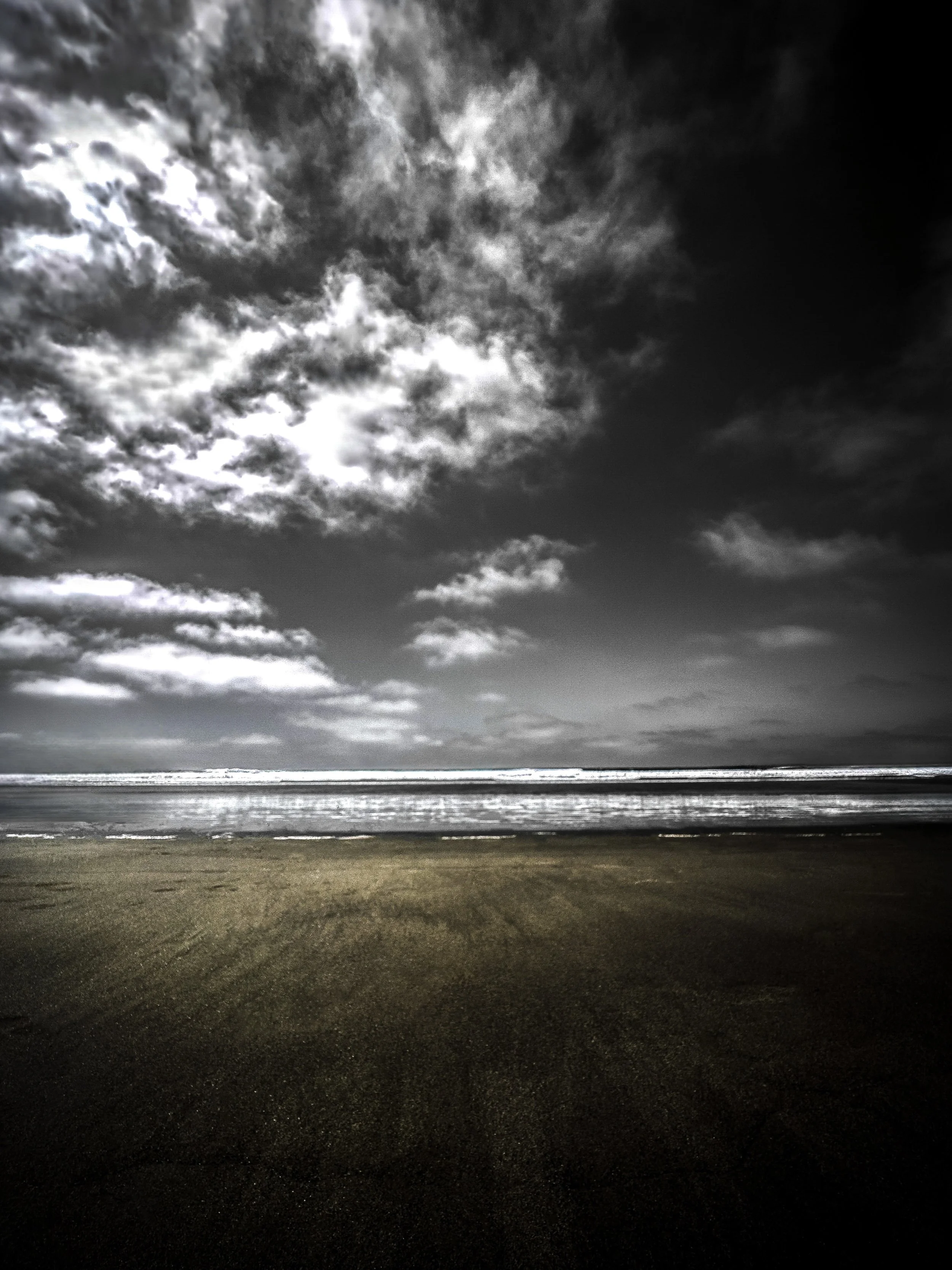 Black and white image of a beach with a dark, cloudy sky and waves crashing onto the shore.
